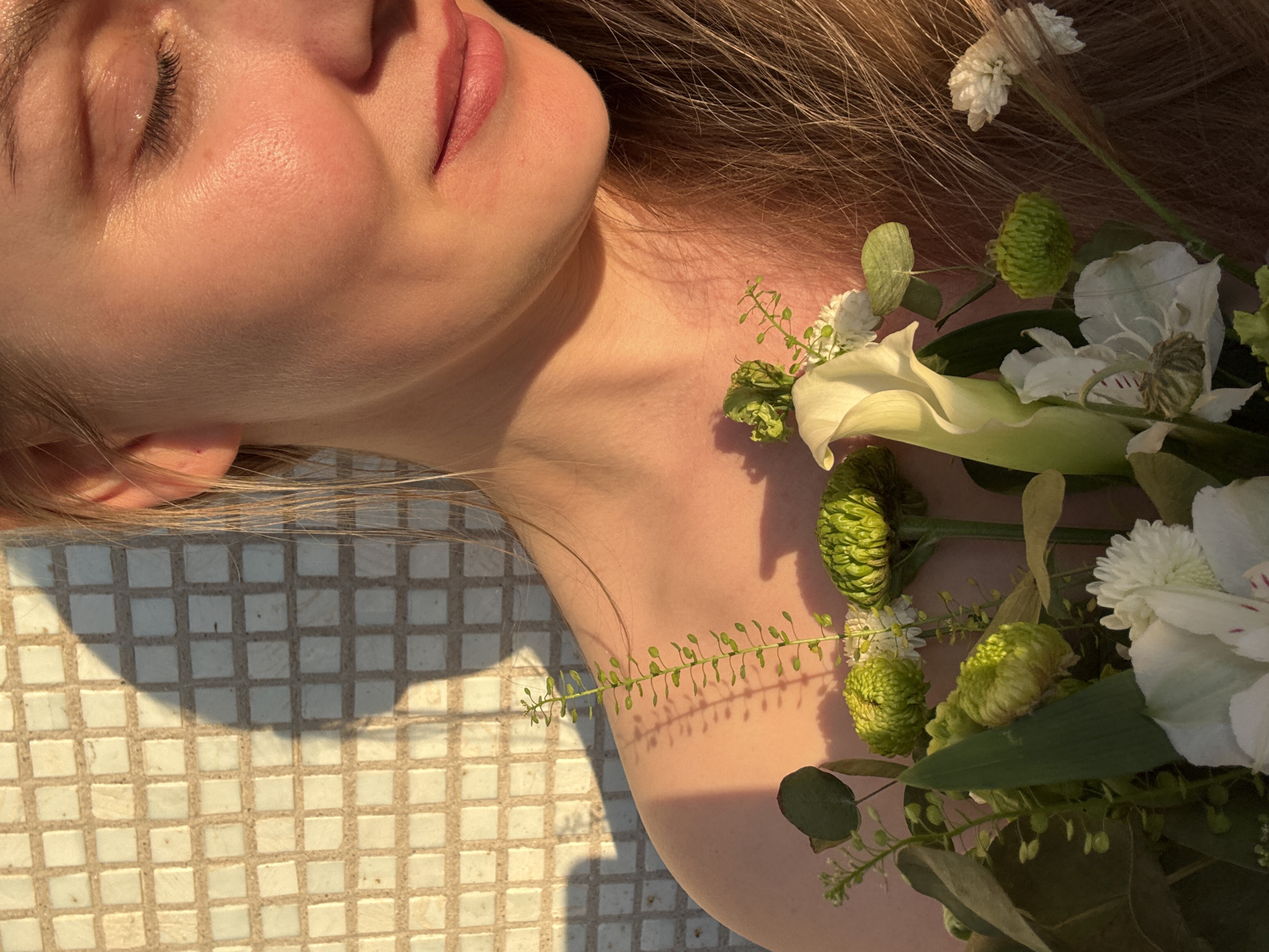 A close-up shot features a light-skinned individual with closed eyes and blonde hair, positioned next to a cluster of white and green flowers. The person's face and upper body are illuminated by strong sunlight, casting distinct shadows. The surface beneath appears to be tiled with small, square tiles. The location or presence of Chaikovsky, Russia cannot be determined from the given visual information.