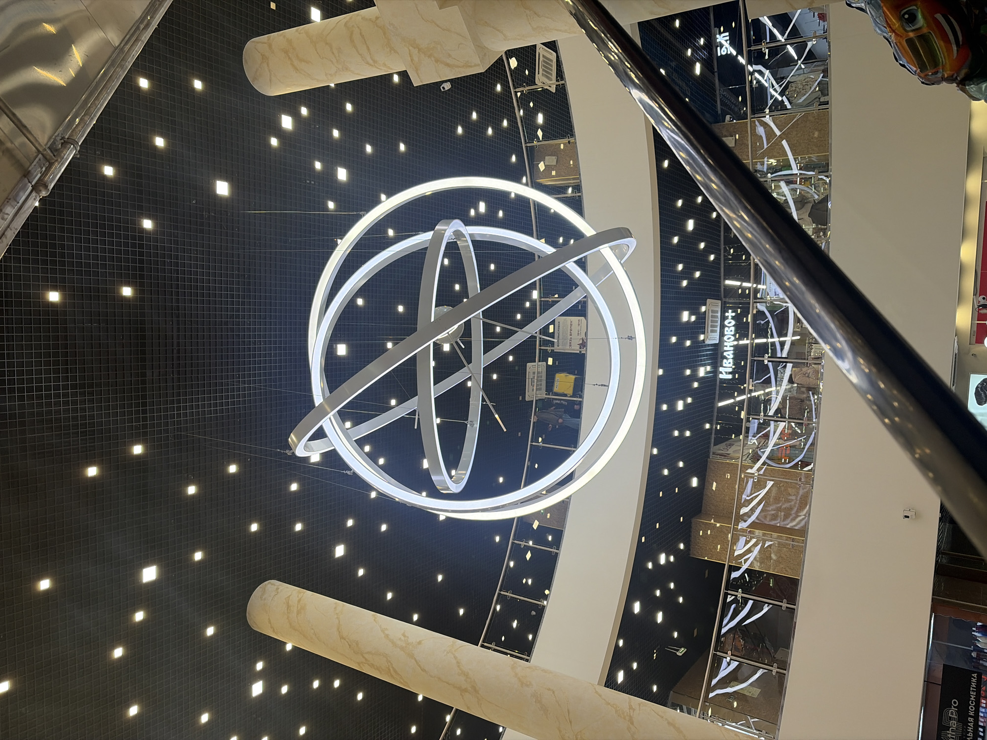 The image is taken indoors, looking up at a decorative lighting fixture in a commercial space, likely a shopping mall in Saint Petersburg, Russia.
The lighting fixture is a series of interconnected white rings that appear to be illuminated with LEDs. The ceiling above is black with a grid pattern and small, square lights. There are also architectural support structures that look like beige-colored pillars, which feature a marbled texture.
In the background, along the right side of the image, there is a multi-story structure that resembles an open-air elevator or escalators. The sides of this structure feature mirrored panels and displays of merchandise. Some signage is visible, including what appears to be the Russian name "ИВАНОВО+". A reflective metal railing is visible in the right foreground, obstructing a clear view of the structure behind.