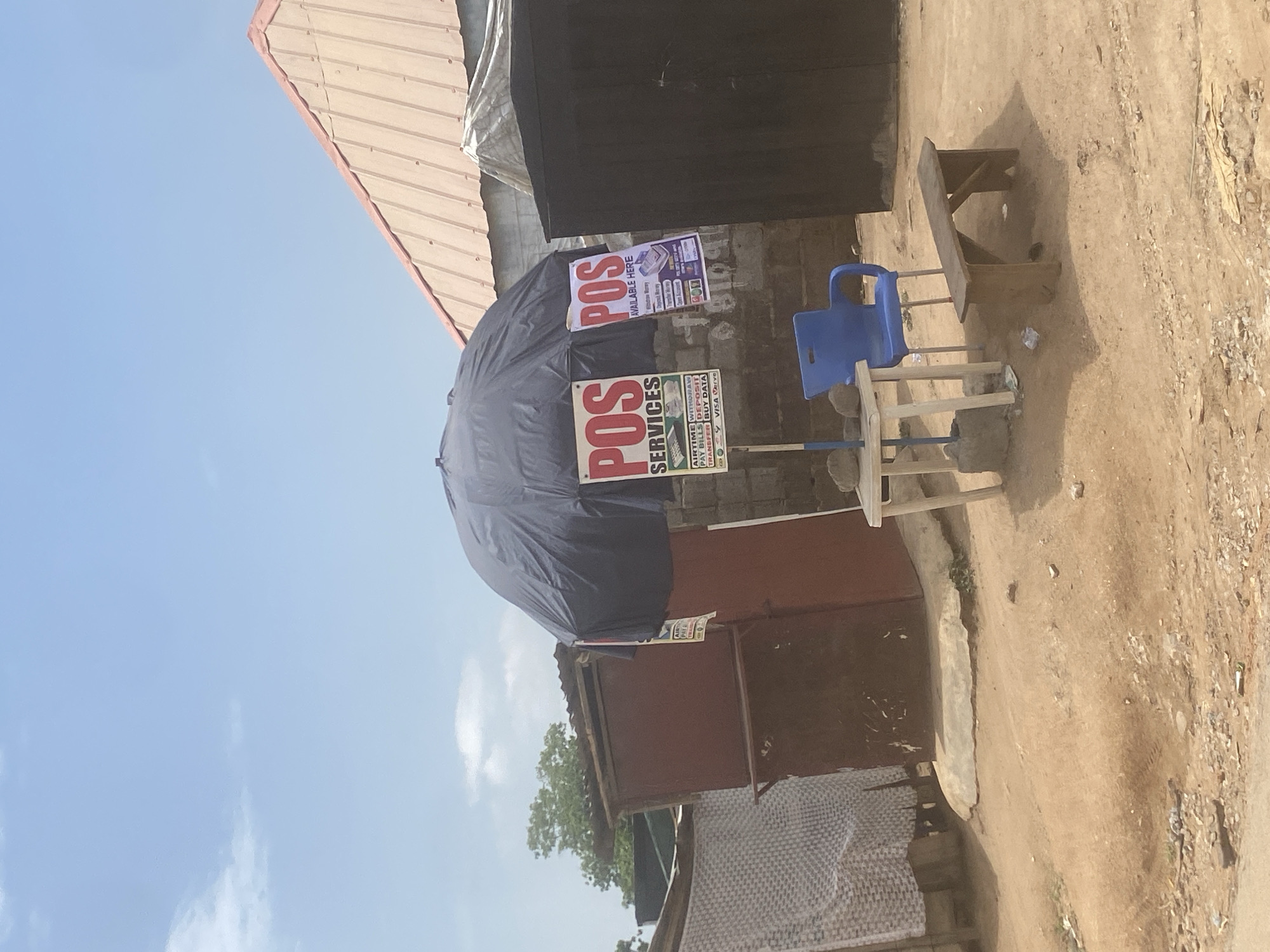 The image shows a street scene in what appears to be a semi-urban or rural setting. There is a simple point-of-sale (POS) business operating under a large black umbrella. The business displays two signs advertising "POS Services" and "POS Available Here." These signs indicate the services offered, such as airtime, withdrawals, bill payments, transfers, and data purchases. The business is equipped with a blue plastic chair and a small wooden table. The surroundings include a dirt path or road, a building with a corrugated metal roof, and other structures that seem to be part of a simple commercial area. It's not possible to determine the exact location in Abuja, Nigeria, from the image alone. There are no people visible in the frame.