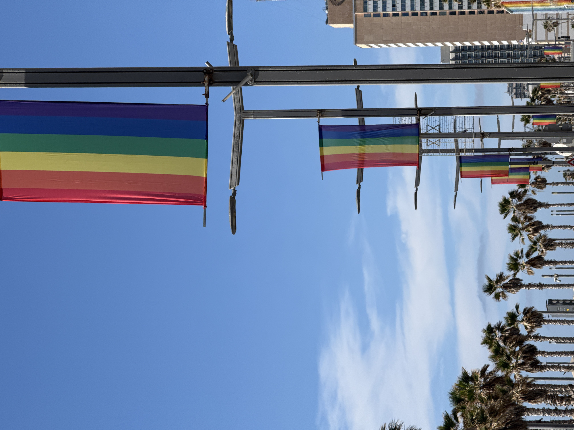 The image shows a row of rainbow flags hanging from metal poles, likely along a street in Tel-Aviv, Israel. The flags are oriented horizontally with the traditional rainbow color sequence (red at the top, violet at the bottom). In the background, there are palm trees and a building. The sky is blue with wispy clouds. The scene is photographed from a low angle, looking upward.