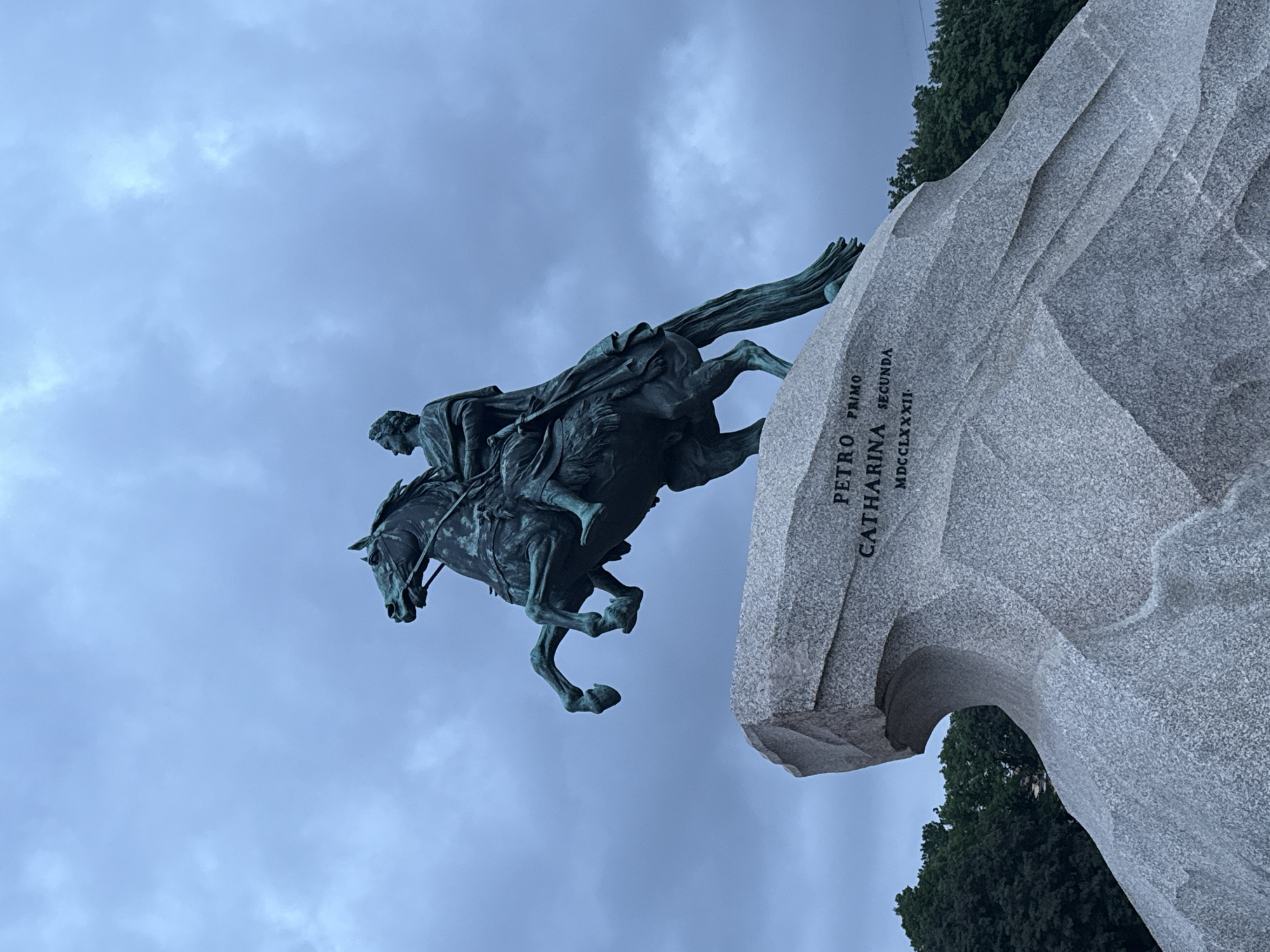 The image shows a bronze equestrian statue of Peter the Great, known as the Bronze Horseman, situated on Senate Square in Saint Petersburg, Russia. The statue depicts Peter the Great on a rearing horse, crushing a serpent beneath its hooves, symbolizing his victory over the Swedes. The statue is mounted on a massive granite boulder known as the Thunder Stone, which was quarried and transported to the site. The sky above is overcast with dramatic clouds, and a line of trees is visible in the background. Inscriptions on the base of the statue read "PETRO PRIMO CATHARINA SECUNDA MDCCLXXXII."