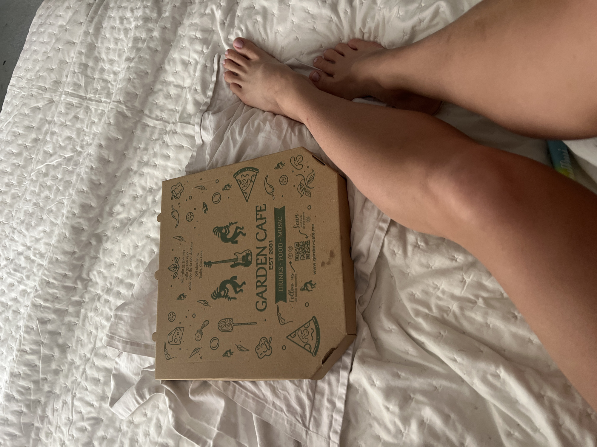 The image shows a pizza box from "Garden Cafe" placed on a white, quilted bedspread next to a person's legs. The pizza box has the cafe's logo, including a guitar, and states "DRINKS FOOD MUSIC EST 2001." The box includes contact information: tel: +382 33 560 293, +382 33 200 090, mob: 067 60 61 60 dostava, XVI ulica br. 23 Budva, Crna Gora and the website www.garden-cafe.me. The legs of an individual are present to the right of the box and feet toward the top of the image.