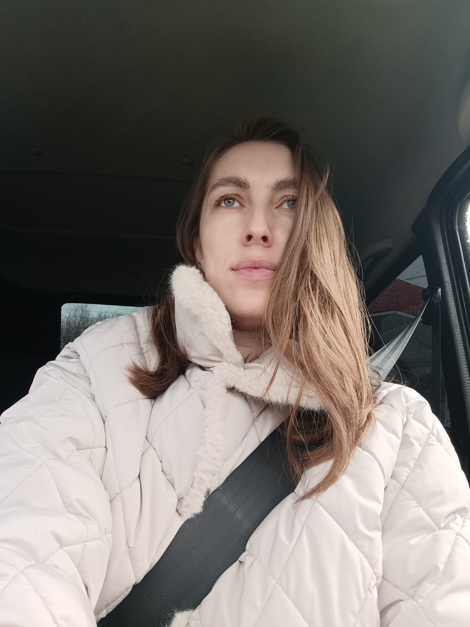 A fair-skinned woman with light brown hair is in a vehicle, wearing a quilted white coat with a faux fur collar and a seatbelt. Her gaze is directed upwards and slightly to the right. Outside the car window, blurred trees and a building with a red roof can be seen. The location is identified as the city of Chaikovsky, Russia.