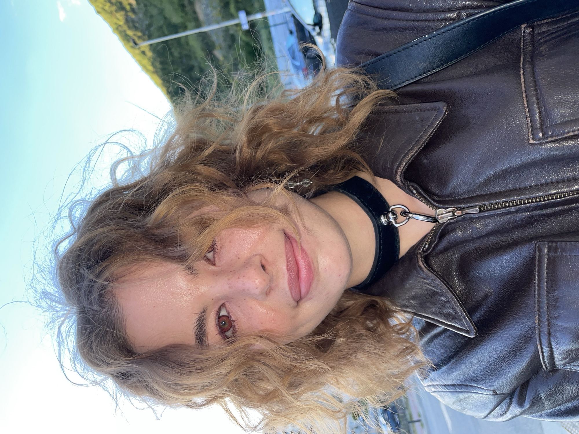 A close-up, tilted portrait features a young woman outdoors in Boreti, Montenegro. Her face, framed by wavy light brown hair, is prominent in the foreground, turned slightly to the right. She has reddish-brown eyes, prominent freckles across her nose and cheeks, and offers a gentle smile with her tongue subtly visible.
She is dressed in a dark brown or black leather jacket with a zippered front and a structured collar. Around her neck, she wears a black choker adorned with a silver D-ring and a small silver charm. A black strap, likely from a bag, is visible over her left shoulder.
The background is softly blurred, revealing a bright blue sky on the left and green foliage or distant hills on the right, typical of an outdoor, sunny setting. The strong, natural light highlights her hair and complexion, suggesting it is daytime. The image appears to be a casual selfie or portrait taken in warm weather. No text is visible.