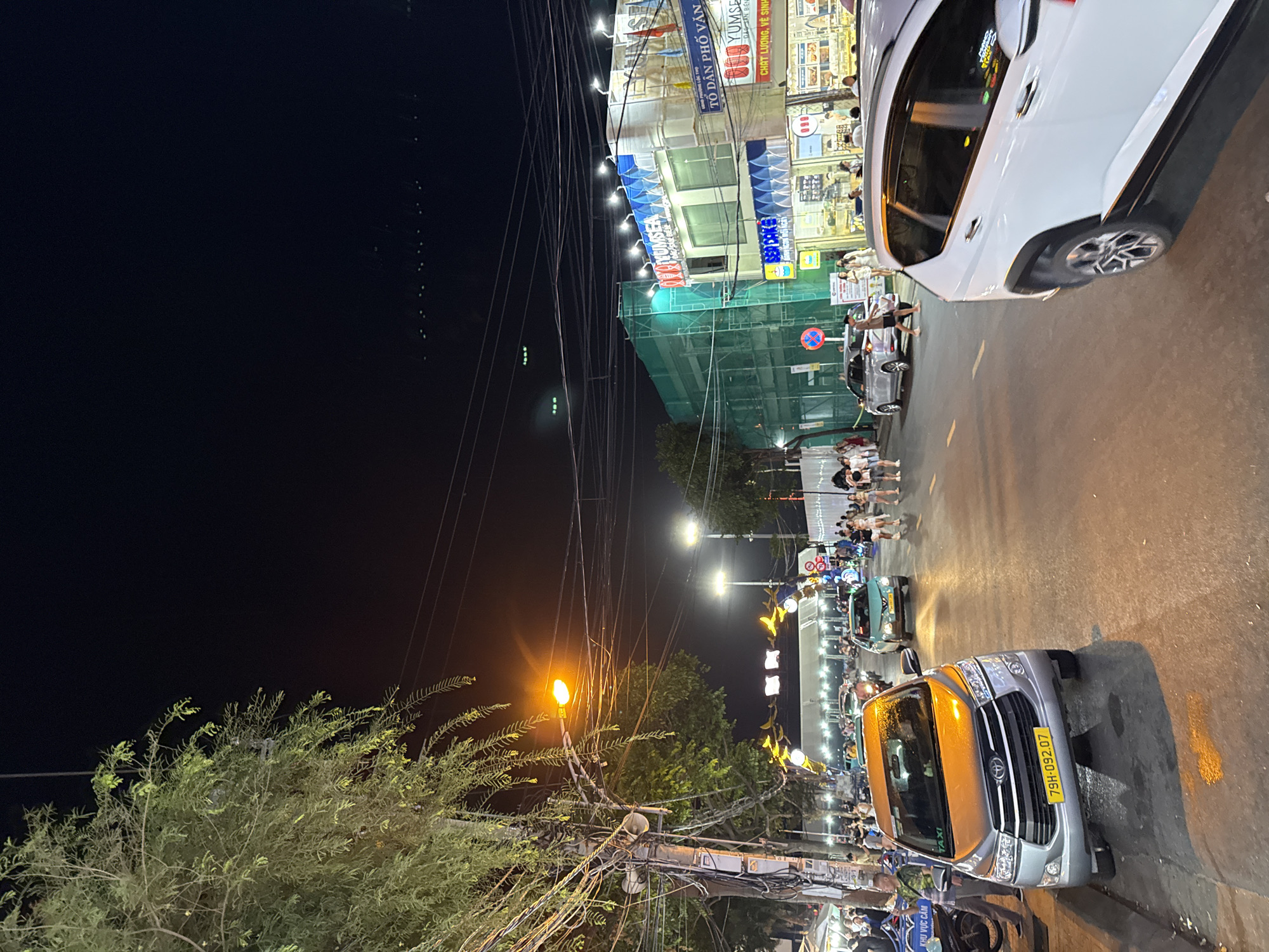This image depicts a street scene at night in Nha Trang, Vietnam, likely a bustling commercial or entertainment district. A bright street light illuminates a road where several cars are present, including a silver Toyota Innova taxi with license plate "79H-092.07" in the foreground. A white SUV is also visible on the right side of the road. The street is lined with buildings adorned with signage, some of which appear to be businesses like "YUMSEA" and "BED CAKE". The buildings are lit up, contributing to the nighttime ambiance. A large structure draped in green netting suggests ongoing construction or renovation. Pedestrians are walking along the sidewalks and crossing the street, some appearing to be tourists or locals enjoying the evening. Overhead, a complex network of electrical wires is strung across poles, a common sight in Vietnamese urban environments. The background shows a dark sky, with distant lights suggesting the presence of more city infrastructure. A large tree with lush green foliage is prominently featured on the left side of the image, its branches partially obscuring the street lights and wires. The overall atmosphere is lively and characteristic of a vibrant night market or city street in Vietnam.