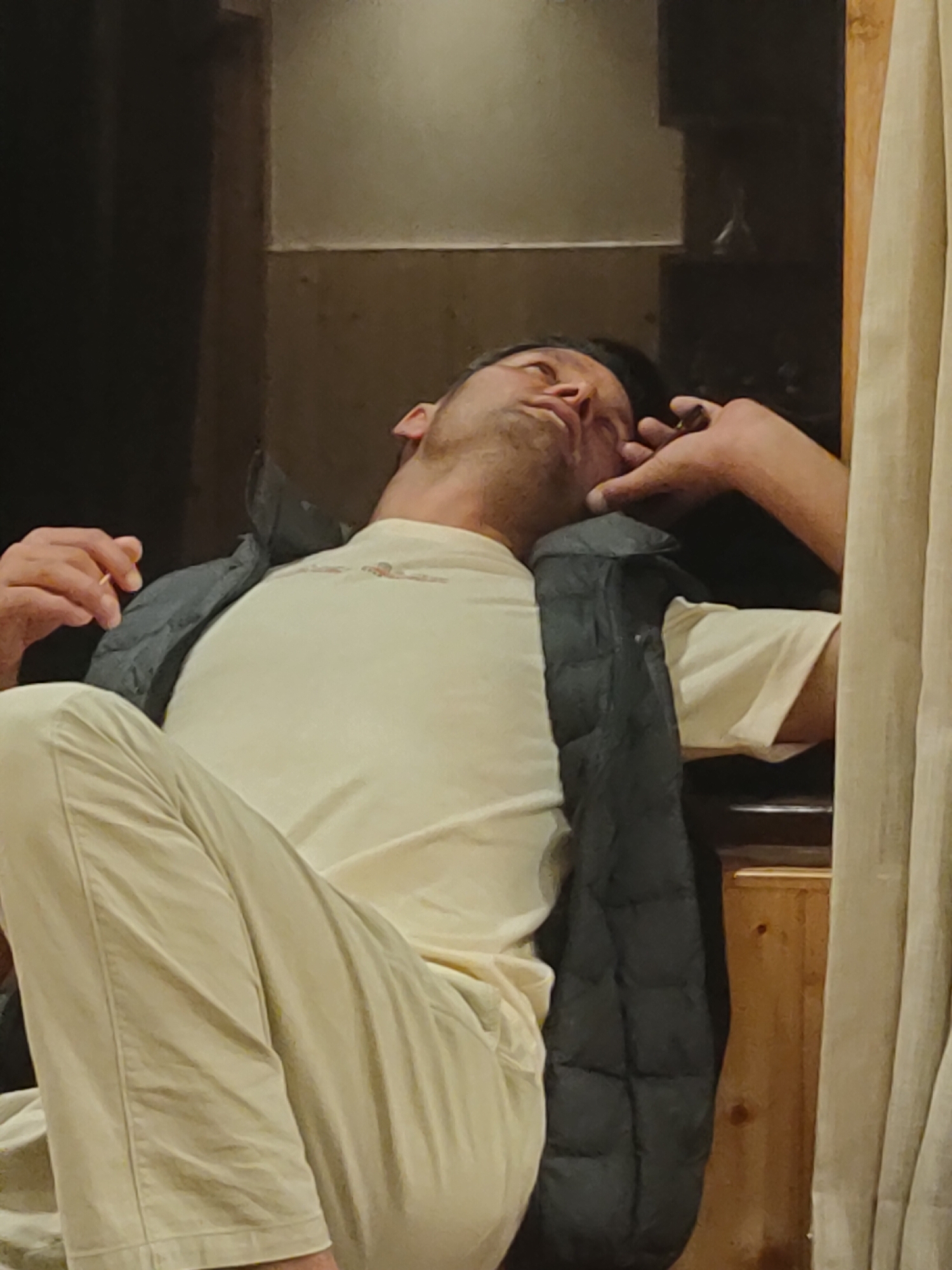 A man is slumped in a chair, head tilted back, eyes closed, and his right hand resting on his face. He is wearing a cream-colored t-shirt and a dark puffer vest over it, with light-colored pants. The background appears to be an indoor setting with wooden accents and beige curtains. There is no indication of the location being in Burwa, India.