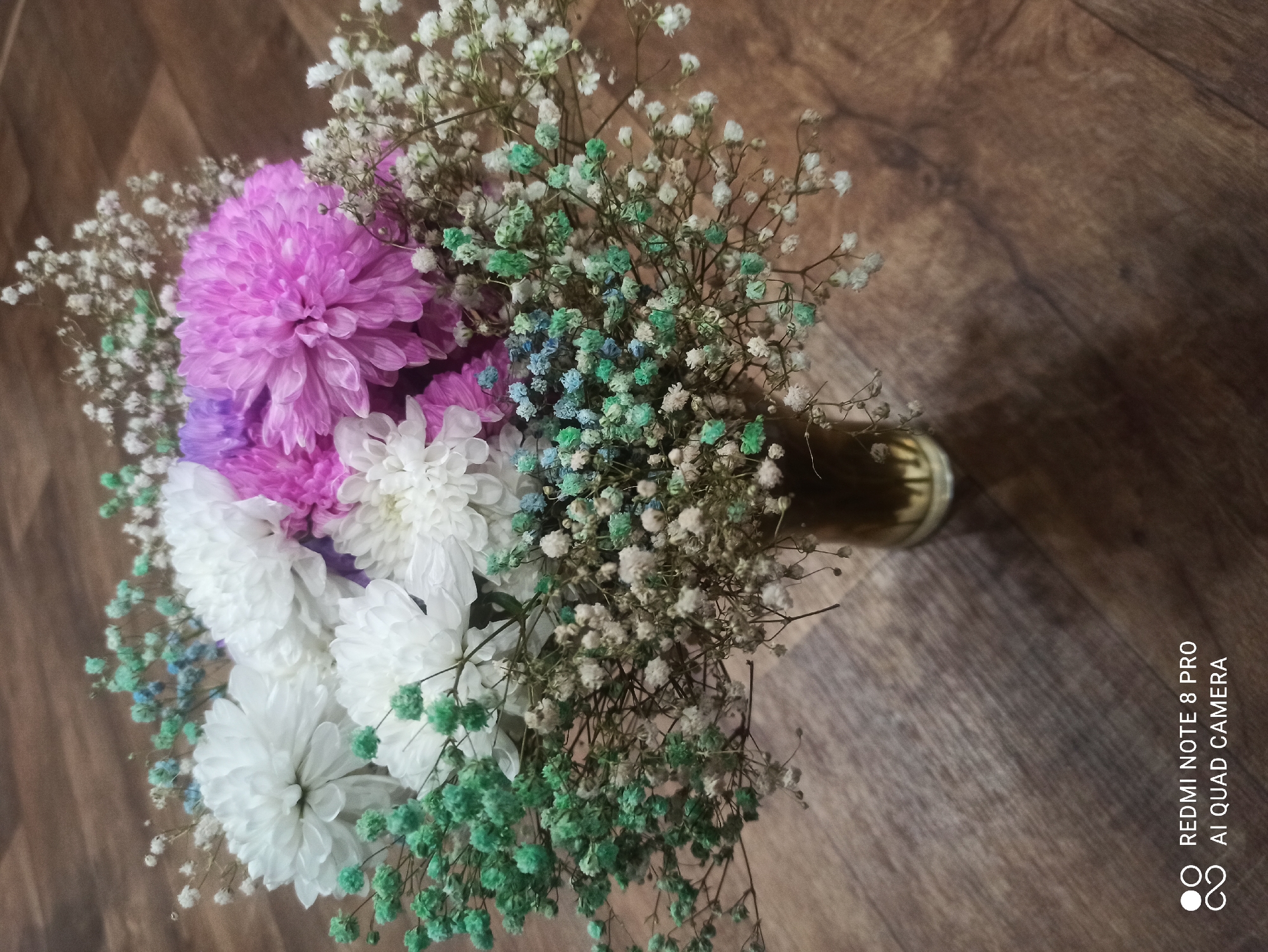 A floral arrangement sits in a glass vase on a wooden surface. The arrangement consists of white chrysanthemums, a purple chrysanthemum, and various small, colorful filler flowers, including green and light blue baby's breath. The background is a brown wooden floor with visible grain. The image is captured indoors, and there is a watermark in the lower left corner indicating "REDMI NOTE 8 PRO AI QUAD CAMERA." There is no indication of the city of Kugesi, Russia, in the image.