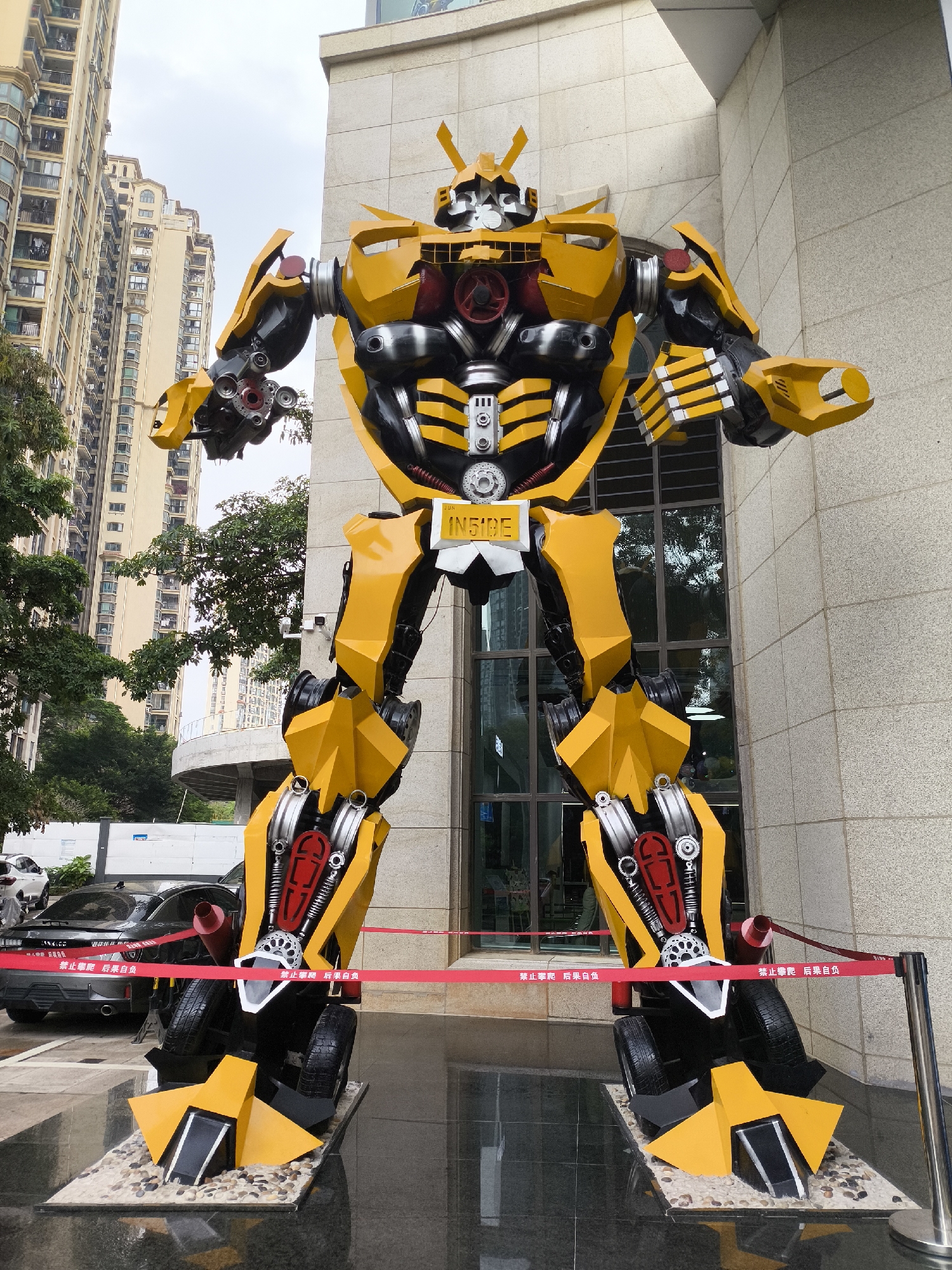 The image shows a large, yellow and black robot statue in front of a building. The robot, which has a sign that reads "INSIDE," is standing on a platform with a red ribbon barrier around it. The statue resembles a Transformer with yellow panels and black and silver mechanical details. In the background, there is a tall, multi-story apartment building and some trees. A black car is parked behind the robot. The image appears to be taken in the Longgang District of China. The sky is overcast. The scene has a combination of architectural and natural elements with an emphasis on the robot sculpture.
