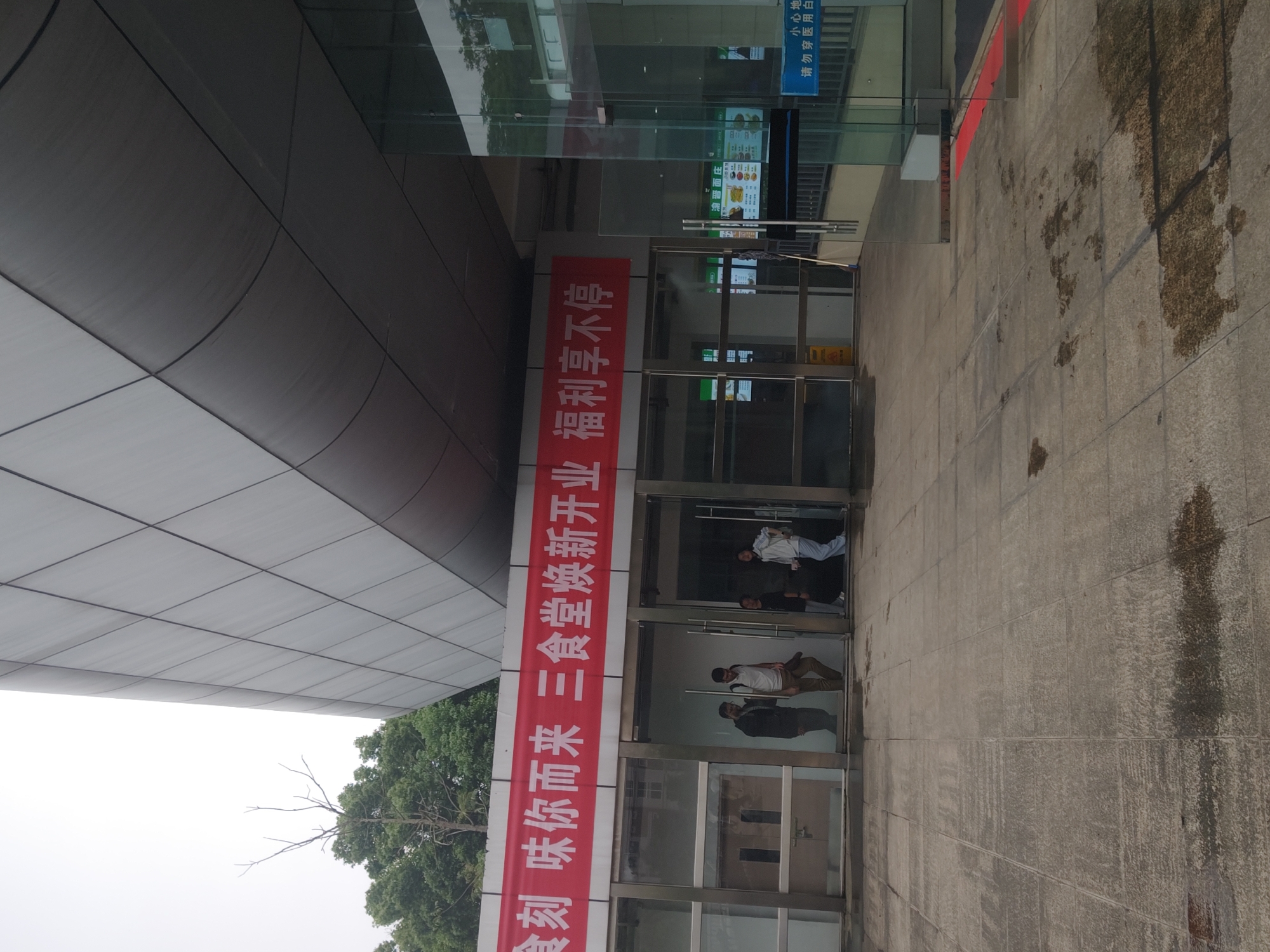 The image shows the entrance to a cafeteria, possibly in Chongqing High-tech Zone, China. A red banner hangs beside the glass doors, featuring Chinese characters vertically aligned, advertising the re-opening of a "three-dining hall" with benefits. Through the glass doors, several people are visible inside the building. The building's exterior is made of grey and white tiles and has some trees visible in the background. The ground leading up to the entrance is tiled and stained. A smaller sign with Chinese writing is visible above the entrance, on the right side of the image.