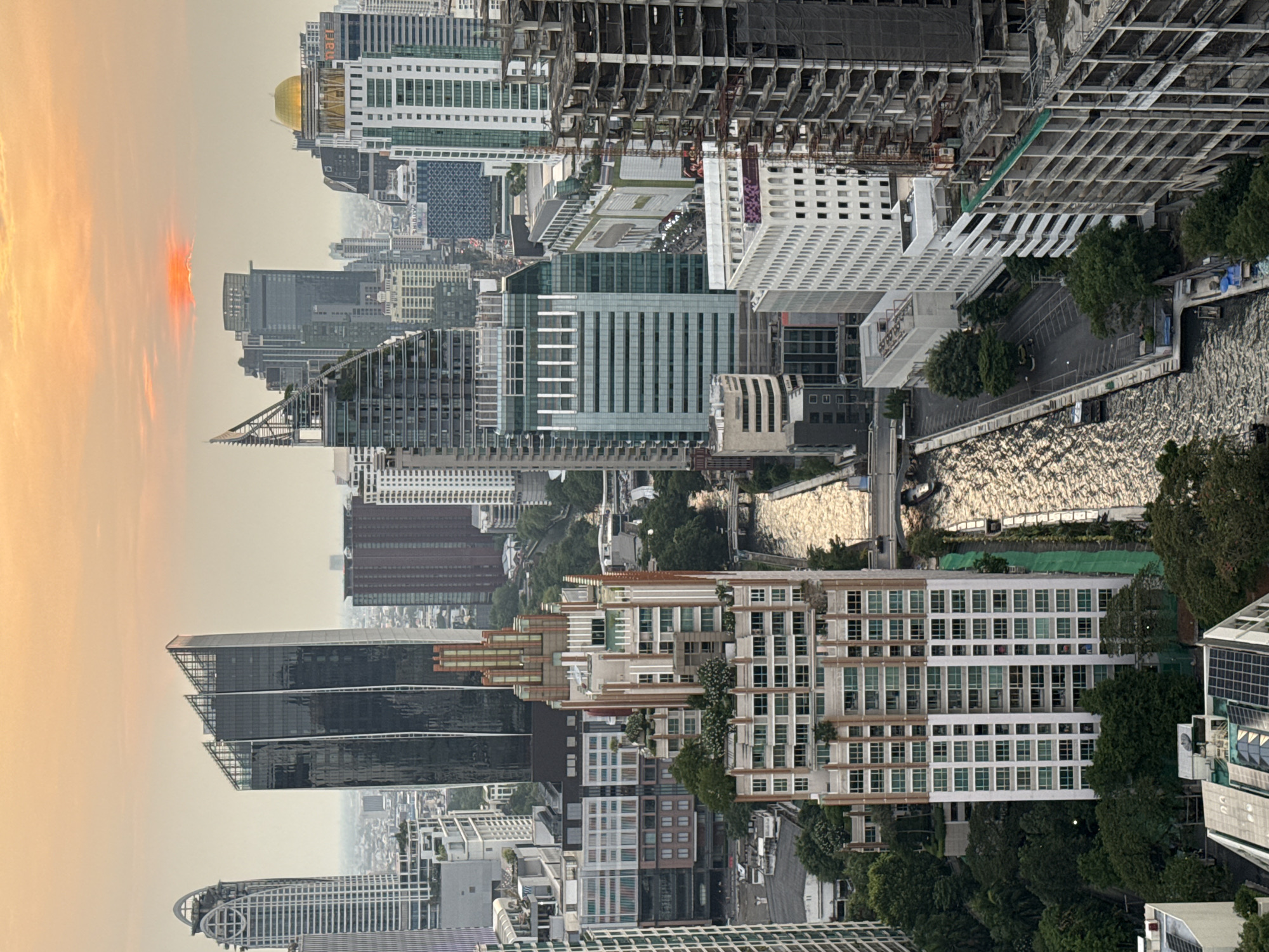 An aerial view depicts Bangkok, Thailand, at dusk. High-rise buildings dominate the scene, showcasing modern architecture mixed with structures under construction. A river bisects the urban landscape, reflecting the ambient light. The sky displays a gradient of colors, transitioning from orange near the horizon to a muted gray above. Construction cranes punctuate the skyline, indicating ongoing development. The image conveys a sense of urban density and growth against the backdrop of a setting sun.