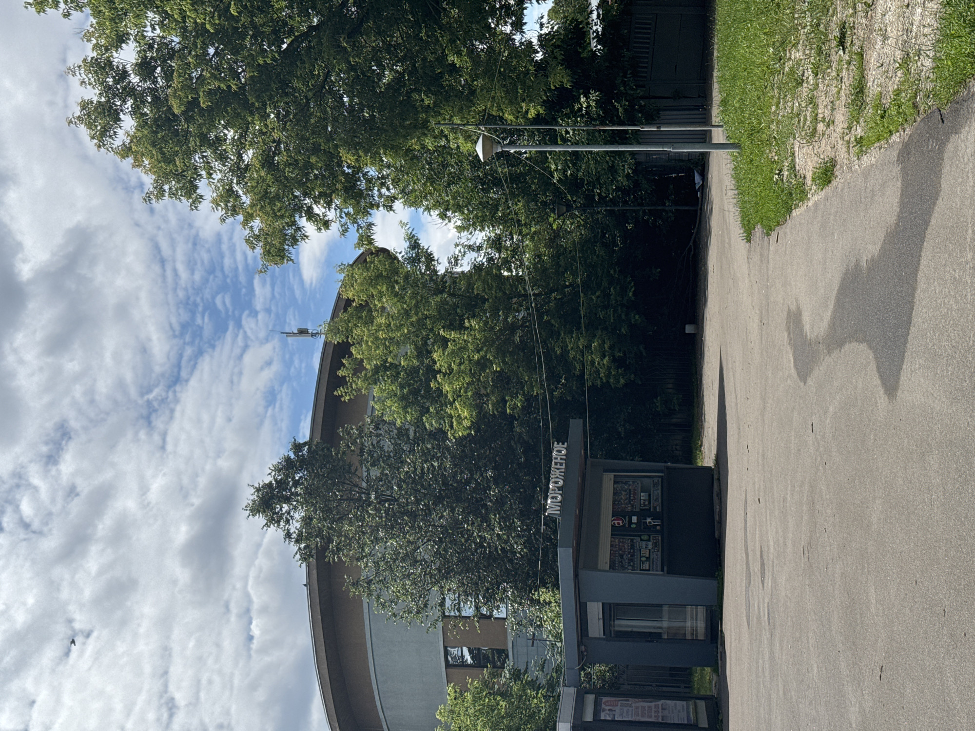 The image depicts an outdoor scene with a circular building in the background, partially obscured by trees. In the foreground, there is a small kiosk with the Cyrillic text "МОРОЖЕНОЕ" (ice cream) visible on its roof. The kiosk appears to be a commercial establishment. To the right of the kiosk, a modern lamppost stands near a grassy area. A paved pathway curves from the foreground towards the right. The sky is partly cloudy. The presence of the kiosk with "ice cream" signage suggests it could be a vendor located in a park or public space. Based on the signage and architectural style, this scene is likely located in Moscow, Russia.