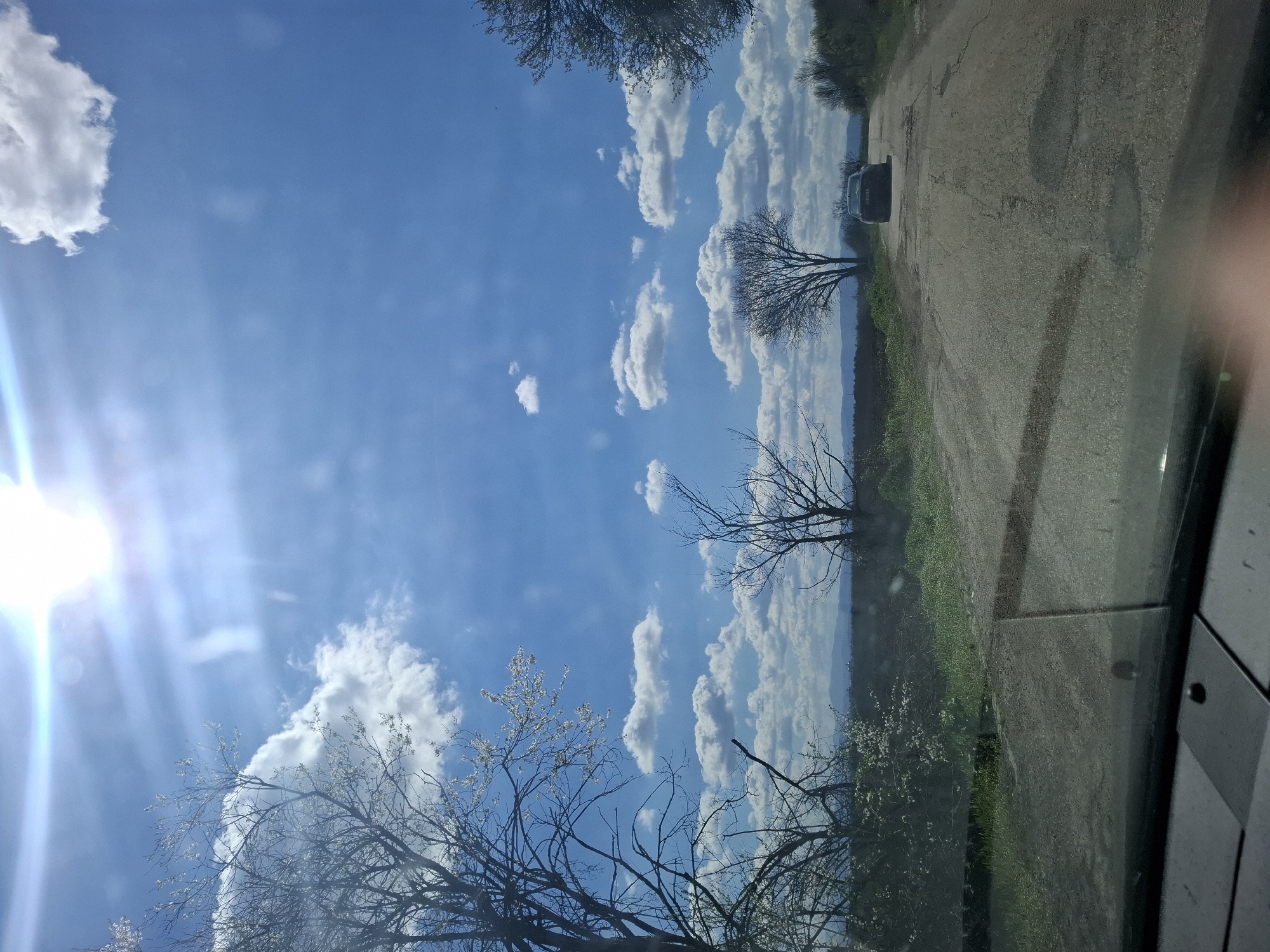 The image shows a view through a car window. The sky is blue with scattered white clouds and the sun shining brightly. Bare trees are visible in the foreground. A road is visible on the right side of the image with a car parked. Green vegetation is present along the side of the road. The location appears to be in Gramada, Bulgaria.