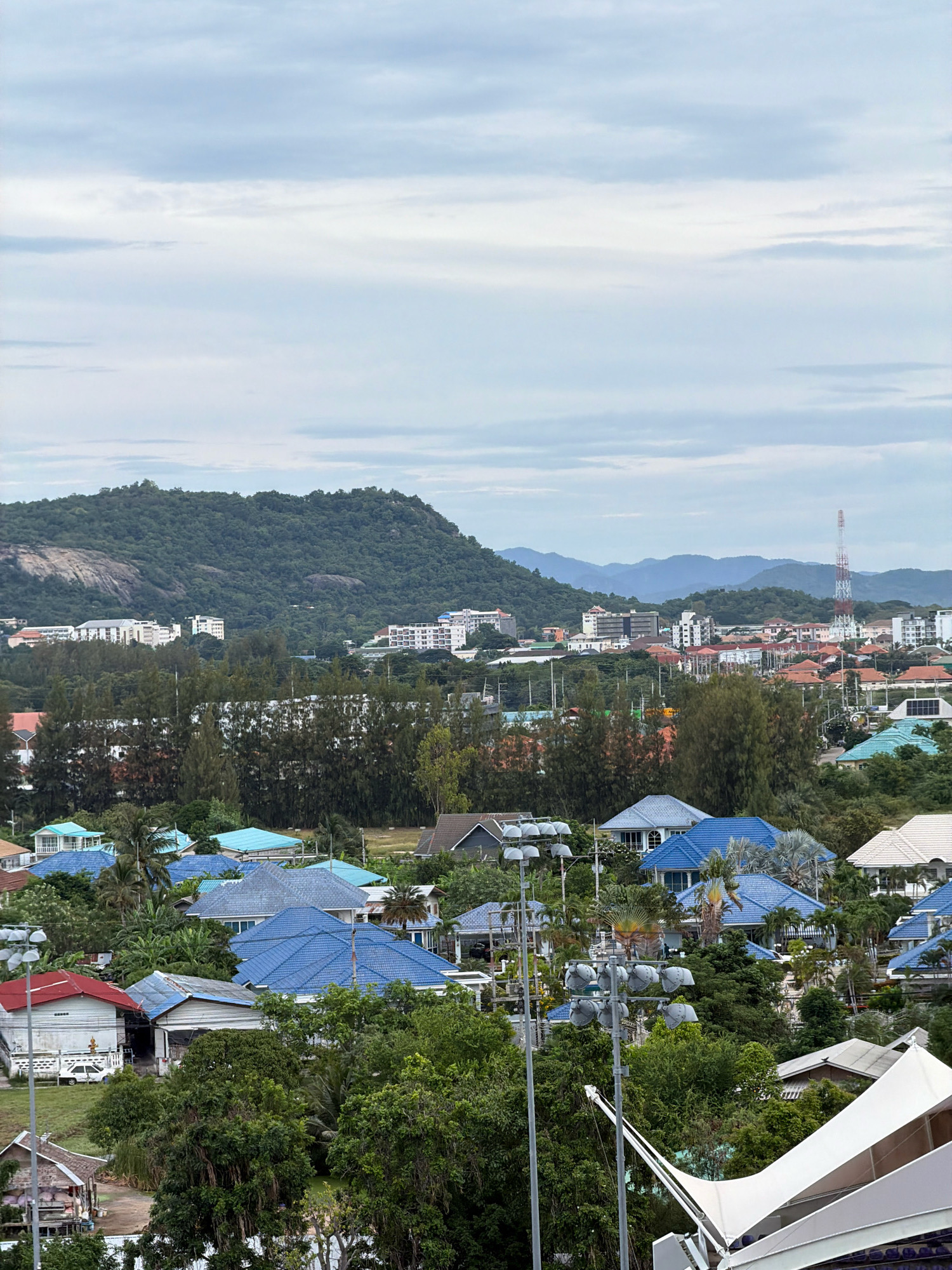 The image captures an aerial view of a residential area in Hua Hin City Municipality, Thailand, dominated by lush greenery and numerous houses with blue and red tiled roofs. A prominent green, tree-covered mountain forms the backdrop, with a cluster of buildings and a tall communication tower visible on its lower slopes. In the foreground, a modern white tent structure with supporting poles is partially visible, suggesting a public space or event venue. The sky is overcast with a light blue and gray cloud cover. The residential architecture features a mix of single-story and multi-story homes, many with spacious yards, interspersed with dense tropical foliage. The scene suggests a suburban or semi-urban setting with a strong natural element.