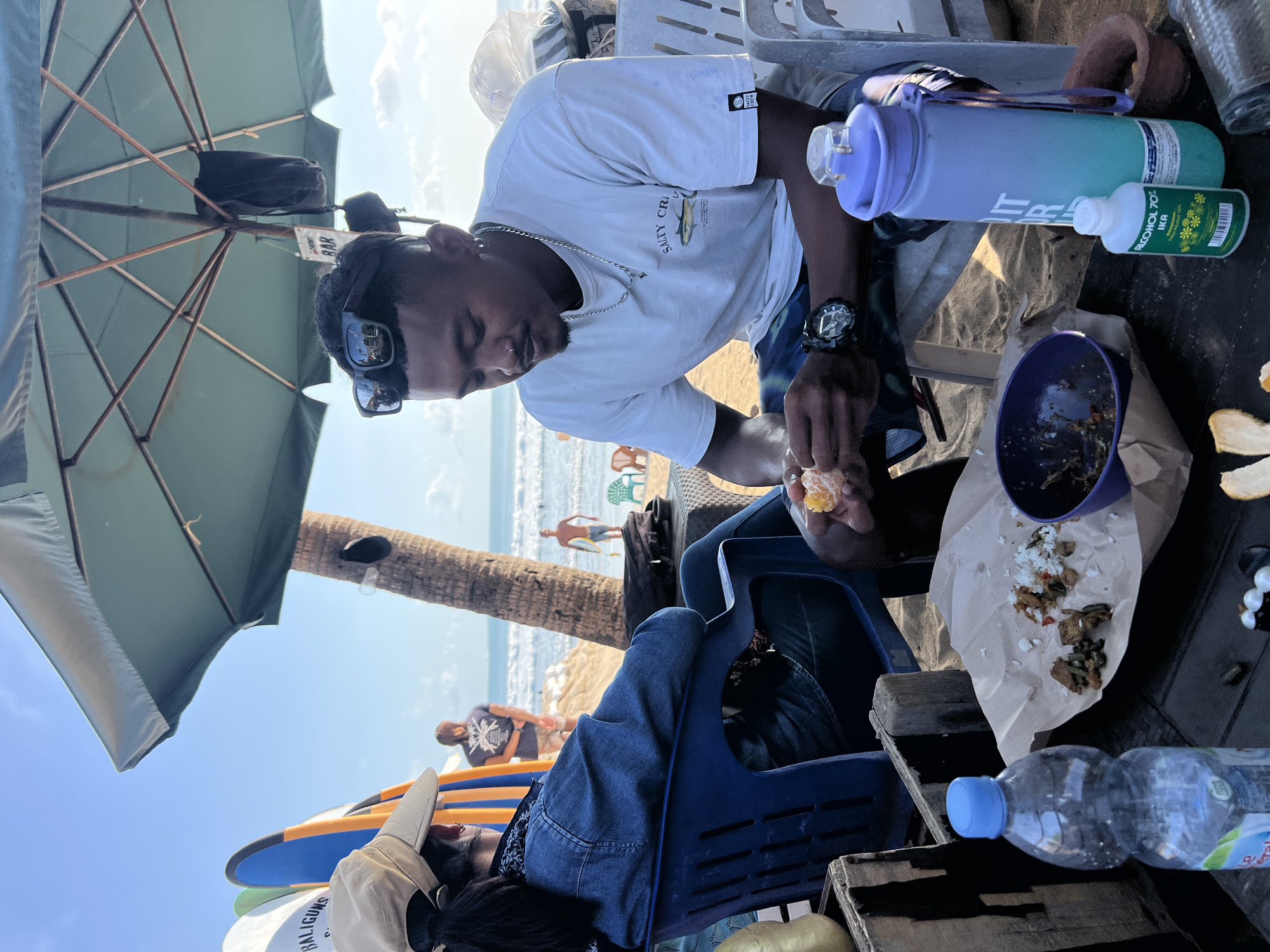 In Kuta, Indonesia, a man and woman are seated at a table on the beach, under a green umbrella. The woman, wearing a tan hat and denim jacket, is partially obscured, facing away from the camera. The man, in a white t-shirt and patterned shorts, is peeling an orange, looking down at his hands. A meal of rice and various food items is spread on brown paper on the table, along with a purple bowl, a water bottle, and a small bottle of alcohol. In the background, the ocean and beach are visible, with surfers in the water and palm trees lining the shore. Several surfboards are propped up to the left of the frame. The overall scene depicts a casual beachside meal in a tropical setting.