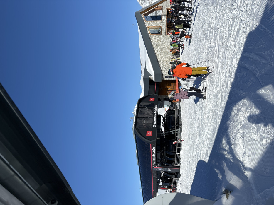 The image depicts a bustling winter scene at a ski resort under a bright, clear blue sky, indicative of excellent weather conditions. The setting is identifiable as Val-d'Isère, France, through the prominent "Télécabine de Solaise" text visible on the black gondola lift cabins.
In the midground, the modern Télécabine de Solaise station is a focal point, with skiers gathering in the snow-covered area around its entrance. Two individuals are notably present on skis near the lift: one wearing a vivid orange jacket and yellow pants, and another in a light purple jacket with black pants. They appear to be preparing to use the lift or have just exited.
To the right, a multi-story building constructed with stone and featuring a grey roof stands amidst the snowy landscape. This structure, likely a mountain restaurant or lodge, is surrounded by numerous skiers and snowboarders, suggesting a popular gathering spot. Their colorful winter clothing adds vibrancy to the snowy environment.
The ground is covered in groomed snow, showing patterns from snow-grooming machines and various ski tracks. Long shadows are cast across the snow, indicating that it is not high noon but likely mid-morning or mid-afternoon, emphasizing the sunny conditions. A dark, angular structure partially frames the scene from the left, possibly part of the lift station or an adjacent building. The overall atmosphere is lively and active, typical of a busy ski day.