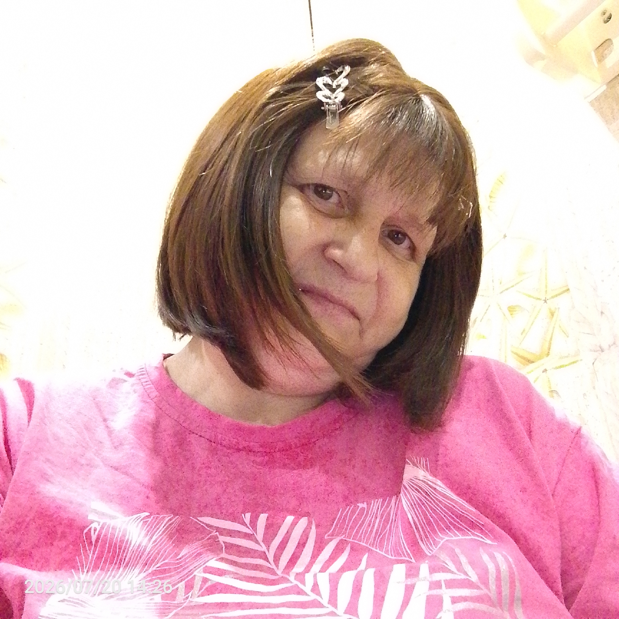 A close-up, low-angle, head-and-shoulders selfie shows a person with brown hair and bangs, wearing a bright pink t-shirt with a white palm leaf pattern. A silver, leaf-shaped hair clip is visible in the person's hair, near the crown. The person's face is turned slightly to the right and their expression appears neutral. The background is out of focus and shows a white wall with some textured, yellow-toned tiling or wallpaper. A timestamp in the bottom left corner reads "2026/07/20 14:26". The image was taken indoors, likely in a bathroom or a room with similar wall coverings.