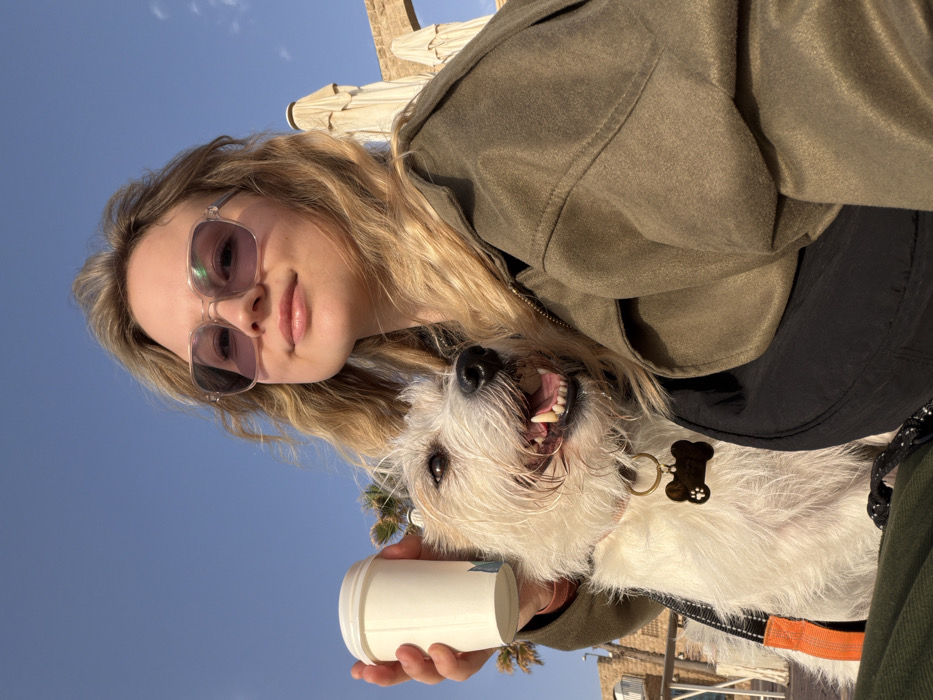 This image depicts a young adult female and a white shaggy-haired terrier-type dog in an outdoor setting at Jaffa Port (Old Jaffa), Tel Aviv, Israel.
The female subject occupies the foreground, positioned slightly right of center, looking towards the camera with a subtle smile. She has long, wavy blonde hair and wears light-tinted square sunglasses. Her attire consists of a dark olive-green zippered jacket over a black garment. Her left hand holds a white disposable coffee cup with a faint blue logo.
To her left, partially obscured by her, is the dog. It is a medium-sized white dog with shaggy fur, wearing a pink collar from which a brass-colored, bone-shaped tag hangs. An orange and black leash or harness strap is visible on its chest. The dog is depicted with its mouth open, panting, revealing its tongue and teeth, suggesting warmth or excitement.
The background features a clear, bright blue sky. To the immediate right, several beige-colored patio umbrellas are visible in a collapsed or folded state. Behind them, ancient light-colored stone structures, including a distinctive archway, are present. Further to the left, palm trees are visible, and in the distance, parts of buildings with white railings and light-colored walls can be seen, consistent with a promenade or port area. The overall scene indicates a sunny day in a historic waterfront district.