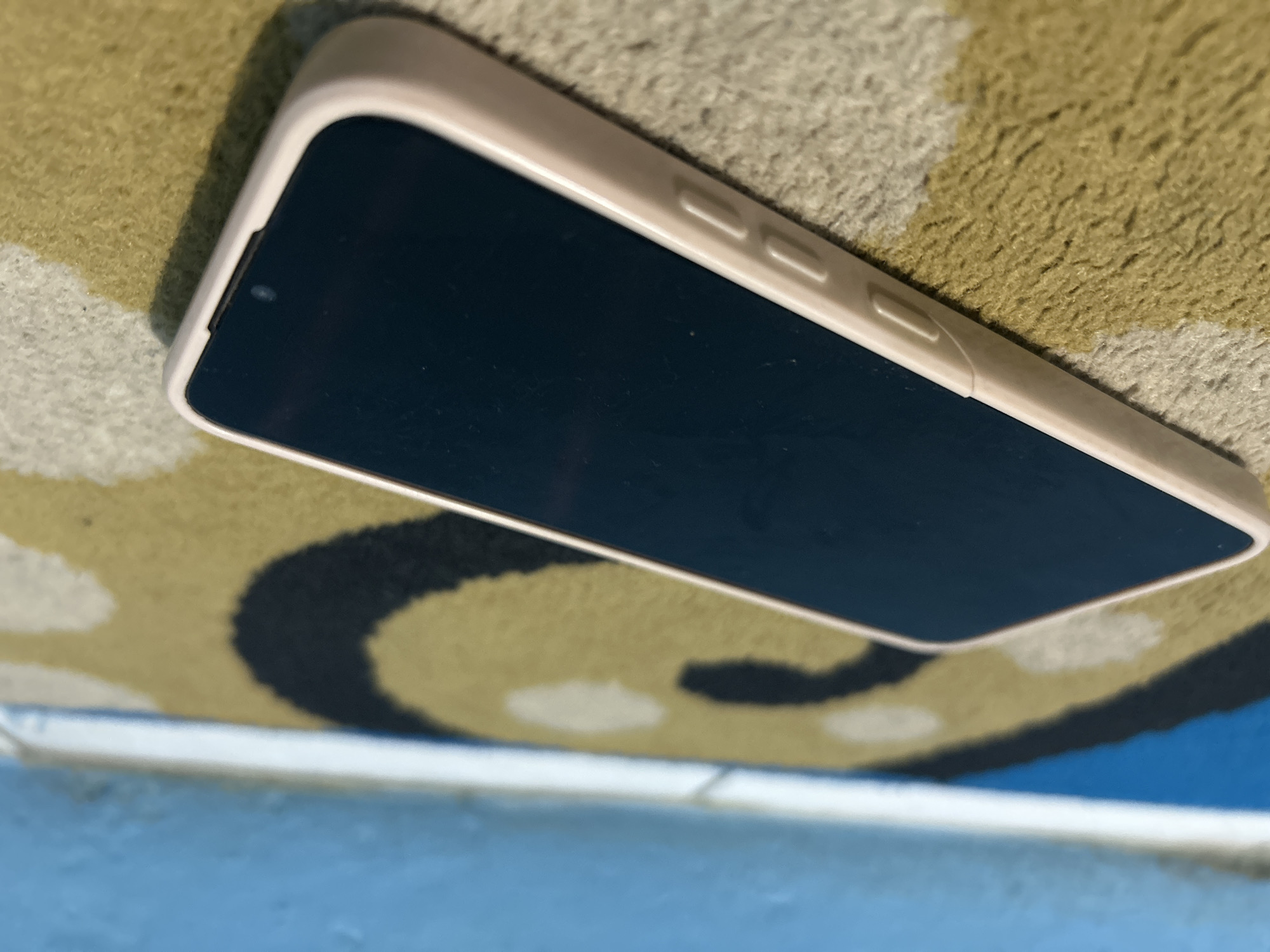 A smartphone with a pink case lies on a textured rug with a black and yellow swirl pattern. The phone's screen is dark. In the background, a blue and white striped surface is visible, which appears to be a wall or furniture. The lighting suggests an indoor setting. The image quality is low, with visible noise and blurriness. The location of Purbalingga, Indonesia is not discernable from the image content.