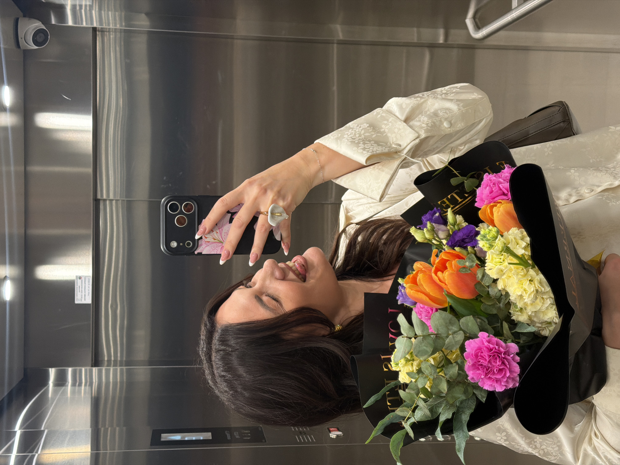 A woman with dark hair is posing for a selfie in an elevator, holding a bouquet of flowers. The elevator has a metallic interior. The flowers include pink carnations and orange tulips. The woman is wearing a light-colored, patterned top and a ring. The city of Belgrade, Serbia is not discernible from the image.