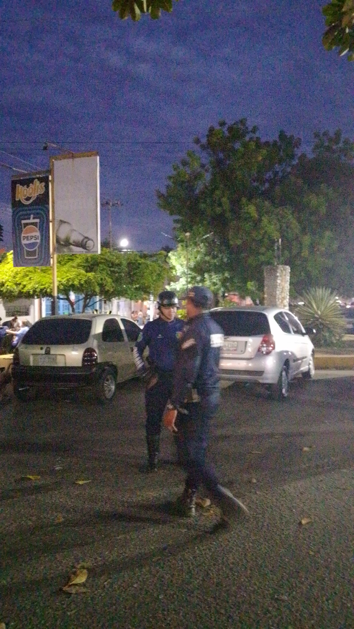 The image shows an outdoor scene at night in Tinaquillo, Venezuela. Two law enforcement officers are standing near parked vehicles. A silver subcompact car is to the left, and a white hatchback is to the right. A tall sign with the “Kool’s” logo and a Pepsi advertisement is visible in the background. The ground appears to be paved with small stones or asphalt. Trees and a streetlight are in the background, contributing to the nighttime environment.