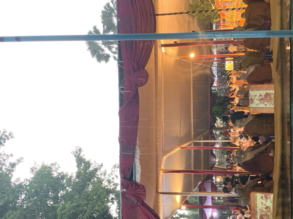 The scene is a gathering of people under a large tent in Mungkid, Indonesia, located at coordinates -7.605, 110.23. The tent is made of white and red fabric, and there are numerous support poles. There is a group of people in orange robes sitting in the front row, facing the camera. Behind them, there are other people seated, mostly in casual clothes. In the background, more people are gathered, and some appear to be dancing or performing. The scene appears to be a gathering or celebration, possibly a religious event. The weather appears to be rainy, as raindrops can be seen falling from the tent's fabric. The sky is overcast, with a white background. There are trees and lush green foliage surrounding the tent. The photo is taken from a low angle, looking up at the scene. The camera is focused on the group of people in orange robes, with the rest of the scene slightly out of focus. The image is slightly dark, but still clear enough to make out the details. The photo was taken at dusk, with the lighting suggesting that the event is taking place at night.