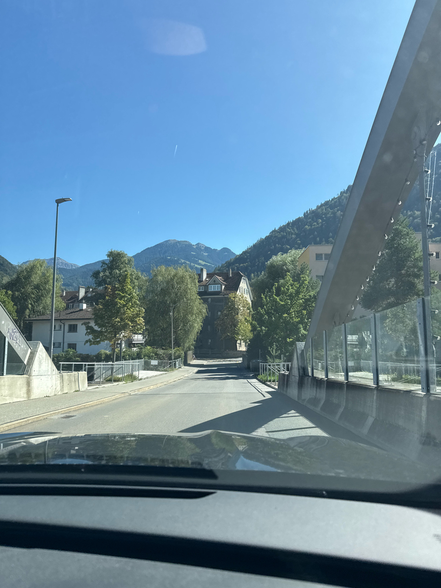 The image depicts a road view from inside a vehicle. The road is paved and curves slightly to the left. On the left side of the road, there is a concrete structure with graffiti and a lamppost. Further down the road, there are several residential buildings and trees. In the background, there are mountains with some greenery. The sky is clear and blue. On the right side of the image, there is a modern bridge with a metal structure and glass panels. The reflection of trees and buildings can be seen on the car's dashboard. The scene appears to be in a town or city situated in a mountainous region.