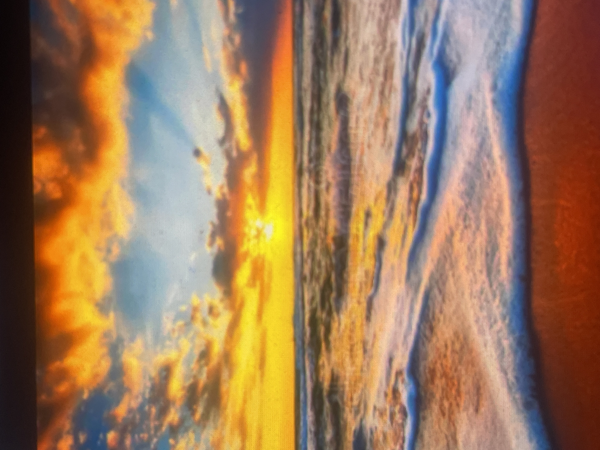 This image depicts a vibrant sunset or sunrise over a coastal landscape in the city of Zelenyi Hai, Ukraine. The upper portion of the scene is dominated by a sky filled with bright orange and yellow clouds, interspersed with areas of blue sky. The sun is visible as a brilliant yellow disc low on the horizon, radiating light across the scene. Below the horizon, a body of water, likely an ocean or sea, extends into the middle ground, reflecting the warm hues of the sky in its surface, appearing predominantly orange and yellow with some darker ripples. Breaking waves are visible across the water, forming white foam as they approach the shore. The immediate foreground reveals a reddish-orange sandy beach, where the waves meet the land, exhibiting white foam and subtle blue tones as the water recedes. No people, structures, or specific objects are present in the image.