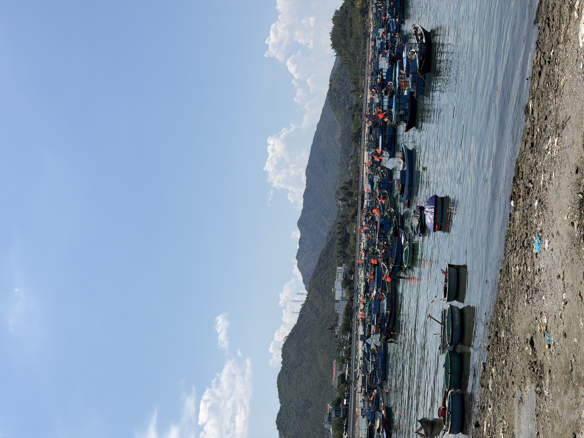 The image shows a harbor in Vĩnh Lương, Vietnam, filled with numerous small fishing boats. The boats are predominantly blue and green, moored densely together. Orange flags are visible on many of the boats. The water is calm, reflecting the sky and the boats. The shoreline is rocky and dirty. In the background, a hill rises with trees covering its side, along with a scattering of buildings. A clear, blue sky with puffy white clouds fills the space above the hill, taking up a large portion of the left side.
