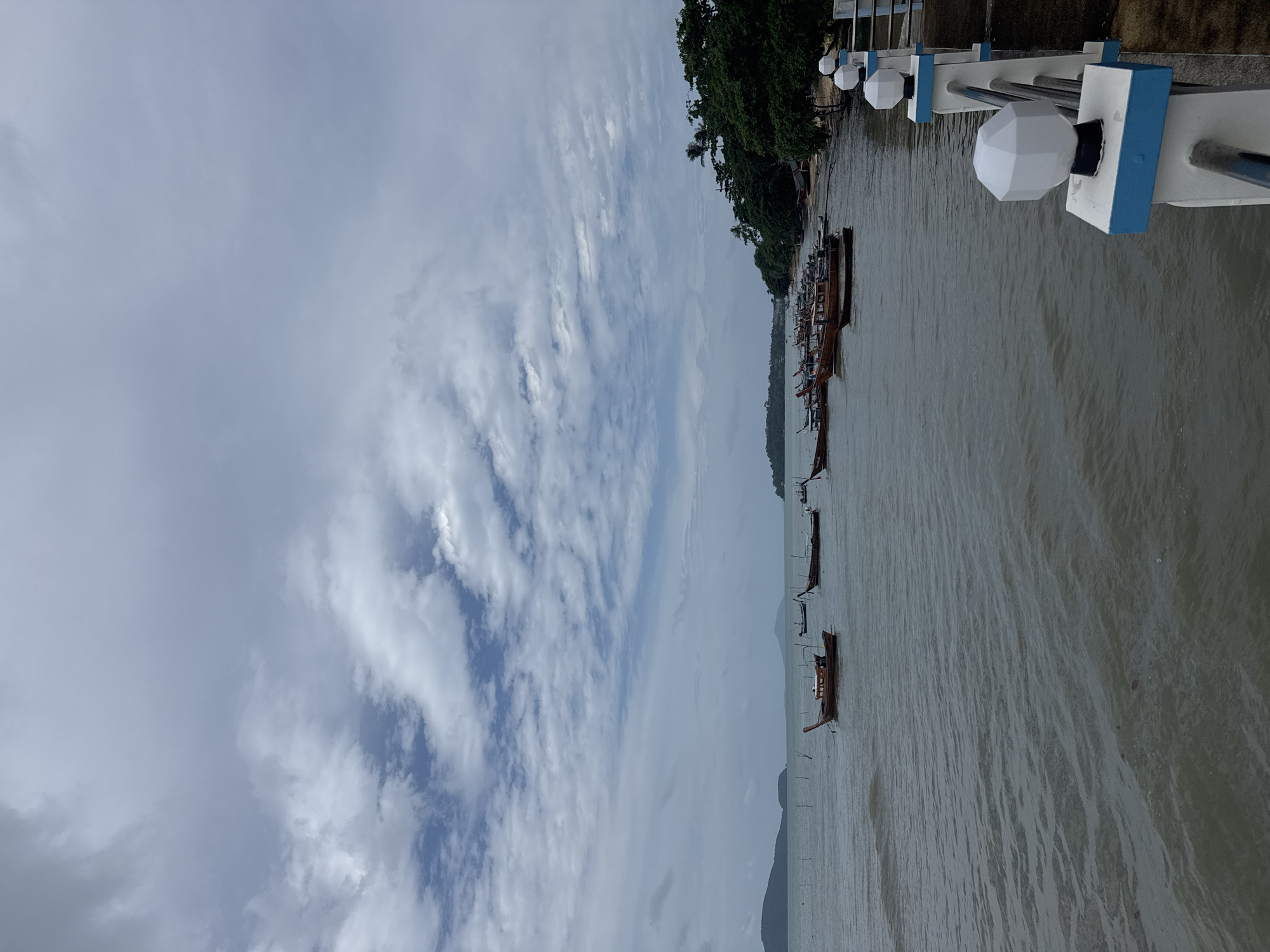 The image depicts a cloudy day at a waterfront in Rawai, Thailand. Several longtail boats are moored in the calm, gray water near a sandy shore lined with lush green trees. In the foreground, a walkway or pier features a white railing with blue accents and hexagonal light fixtures. The horizon shows distant islands and a hazy sky with scattered clouds.