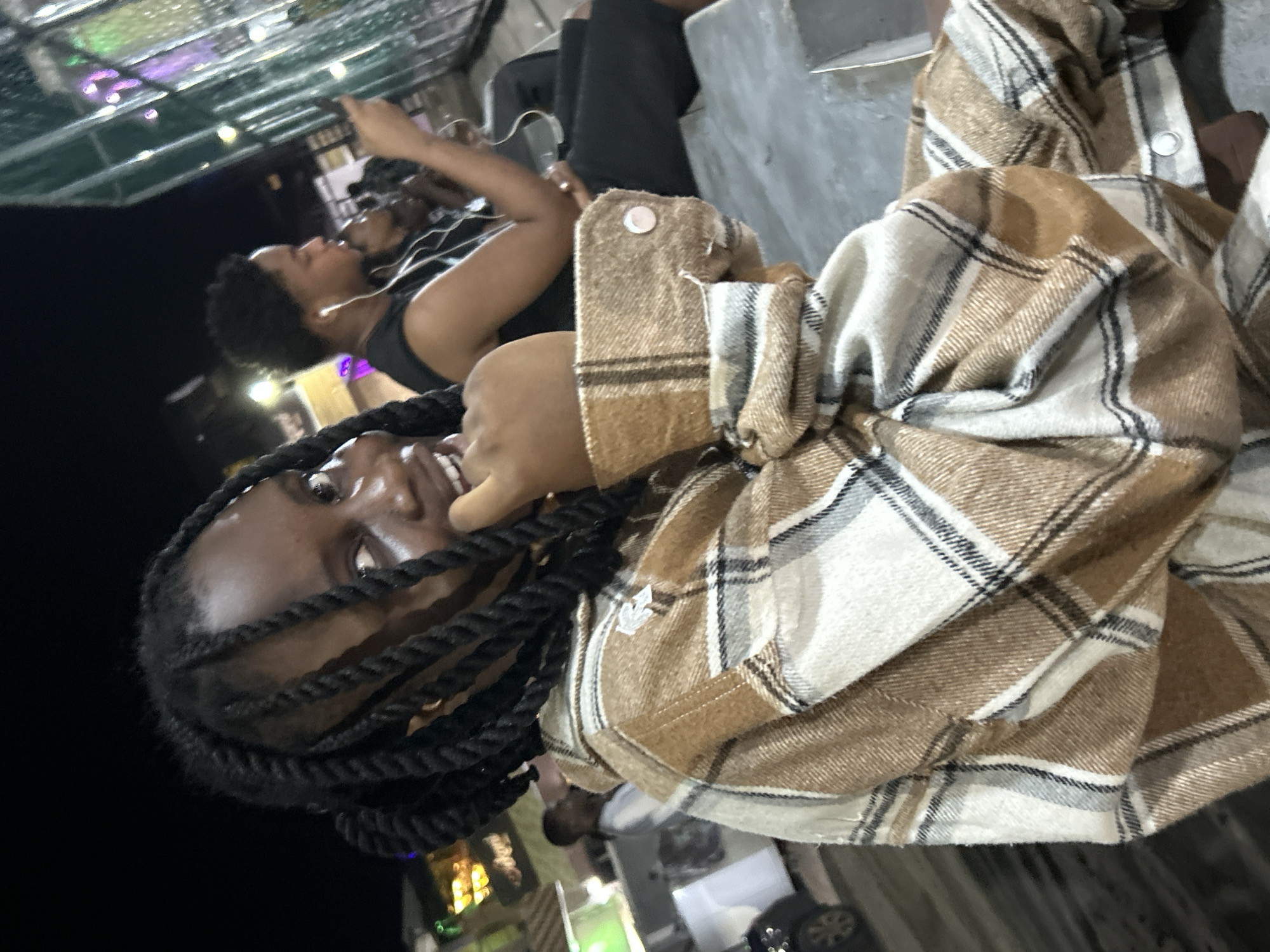The scene depicts an outdoor, urban night environment in Ihiagwa, Nigeria, illuminated by various artificial light sources.
In the foreground, a young individual with dark skin, black twisted hair, and a brown, beige, white, and dark-striped plaid long-sleeved shirt or jacket is positioned. This individual faces the camera directly, displaying a broad smile, with their right hand partially covering their mouth. The cuff of their left sleeve shows a circular, light-colored button or snap fastener.
To the right of the foreground subject, in the mid-ground, another young individual with dark skin and short dark hair is seated in profile, facing left. This person wears a dark, sleeveless top, has white wired earphones in their ears, and is engaged with a black smartphone held in their left hand. Their right arm is bent, resting on their lap. A third, partially obscured individual with dark skin is visible further back and to the right, also appearing to be observing a device.
The subjects are seated on a light-grey, flat surface, possibly a concrete bench or ledge. The background on the right features a tall, green, mesh-like fence or barrier, beyond which various blurred artificial lights, some emitting purple and green hues, are visible. The left background displays blurred, warm-toned lights from distant structures and a partial view of a dark vehicle. The overall illumination is artificial and concentrated, leaving much of the deep background in shadow.