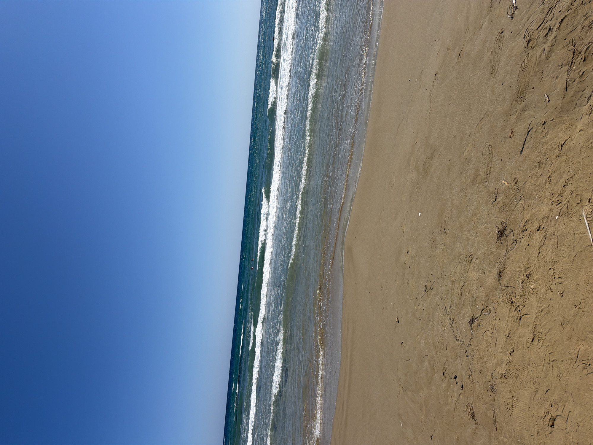 A wide-angle, low-angle shot depicts a sandy beach in Georgioupoli, Greece, under a clear blue sky. The foreground is dominated by sand, showing footprints and small pieces of debris. The middle ground features the gentle turquoise waves of the sea as they approach the shore, creating white foam. In the distance, several figures are visible swimming or wading in the water. The horizon line is sharply defined, separating the sea from the vast expanse of the sky. The overall scene conveys a serene, sun-drenched beach environment.