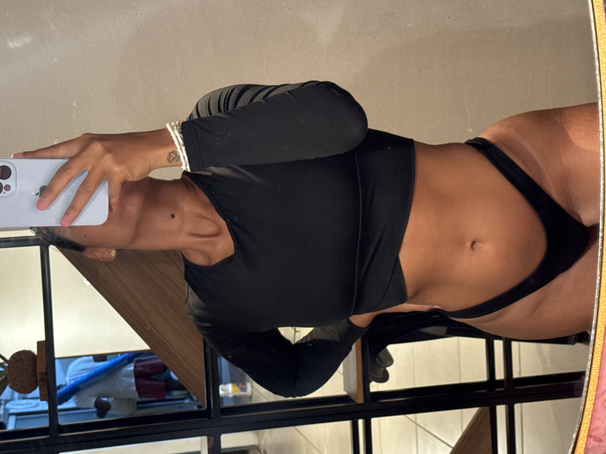 A medium shot depicts a woman in front of a mirror. She is holding a cell phone to take a picture of herself. She is wearing a black top with long sleeves and black underwear. She has a tattoo on her left wrist and a gold ring on her finger. In the mirror's reflection, one can see a portion of a room including a plant and a person using a treadmill. Based on the style of building and surroundings, the image may have been taken in Armação Dos Búzios, Brazil.