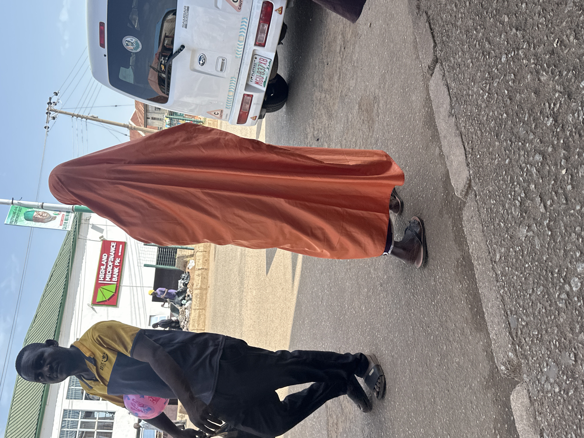 In the city of Bukuru, Nigeria, a man wearing a blue and yellow shirt and black pants stands to the left, holding a pink balloon. Beside him, a person in an orange hijab walks away from the camera. A white van with a license plate that reads "MGU-837XB" is parked on the right. The background shows a building with a sign for "HIGHLAND MICROFINANCE BANK PIC" and a green roof. There are other people and a pole with wires in the distance. The scene appears to be a street.