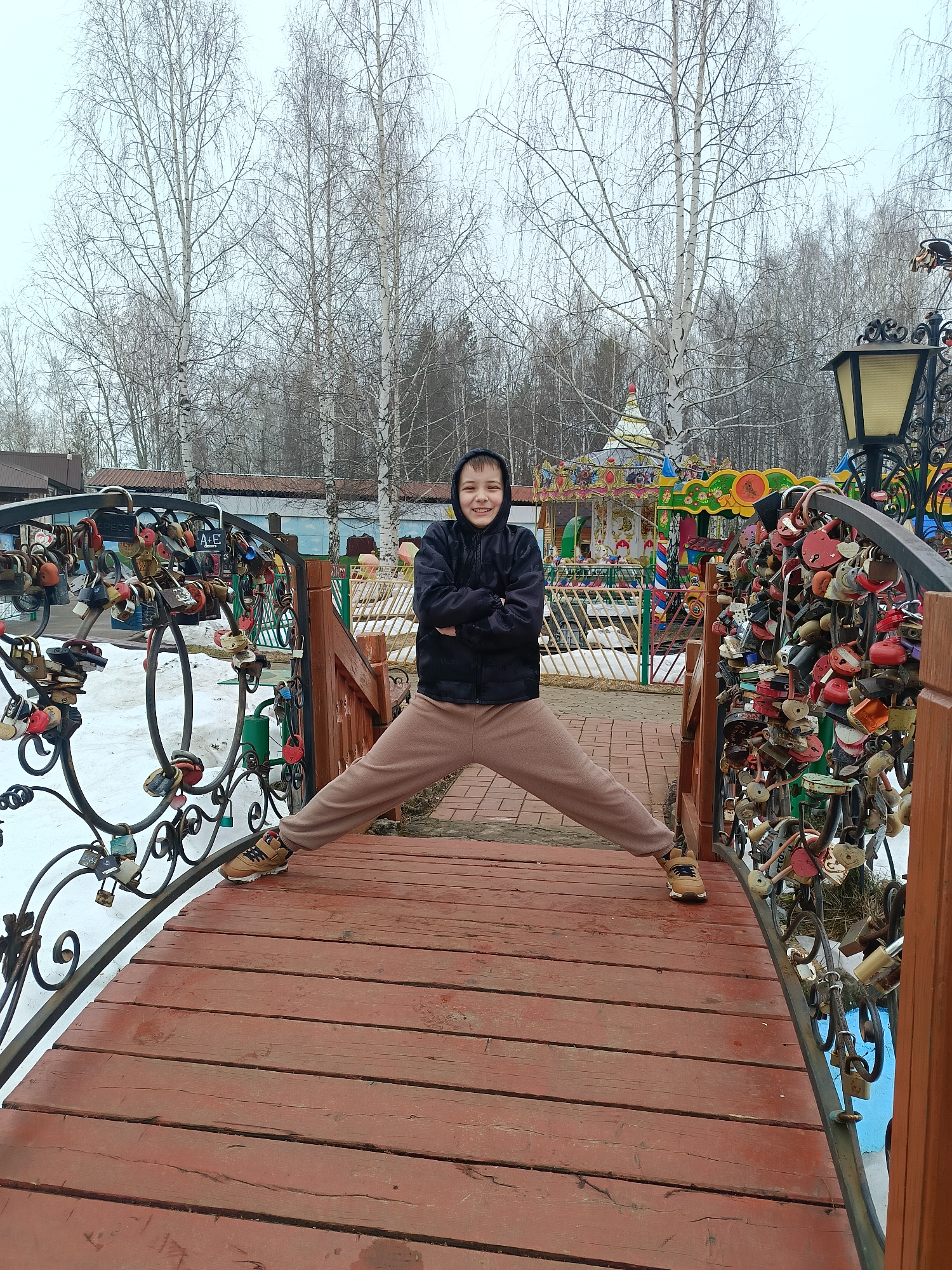 A young person with short brown hair wearing a black hooded jacket and brown pants stands on a red wooden bridge with legs spread apart, arms crossed, and smiling. The bridge's railings are adorned with numerous padlocks. The background features bare birch trees and a carousel. The location is a pedestrian bridge in Chaikovsky, Russia. Patches of snow are visible near the bridge. A vintage-style lamp post is also present.
