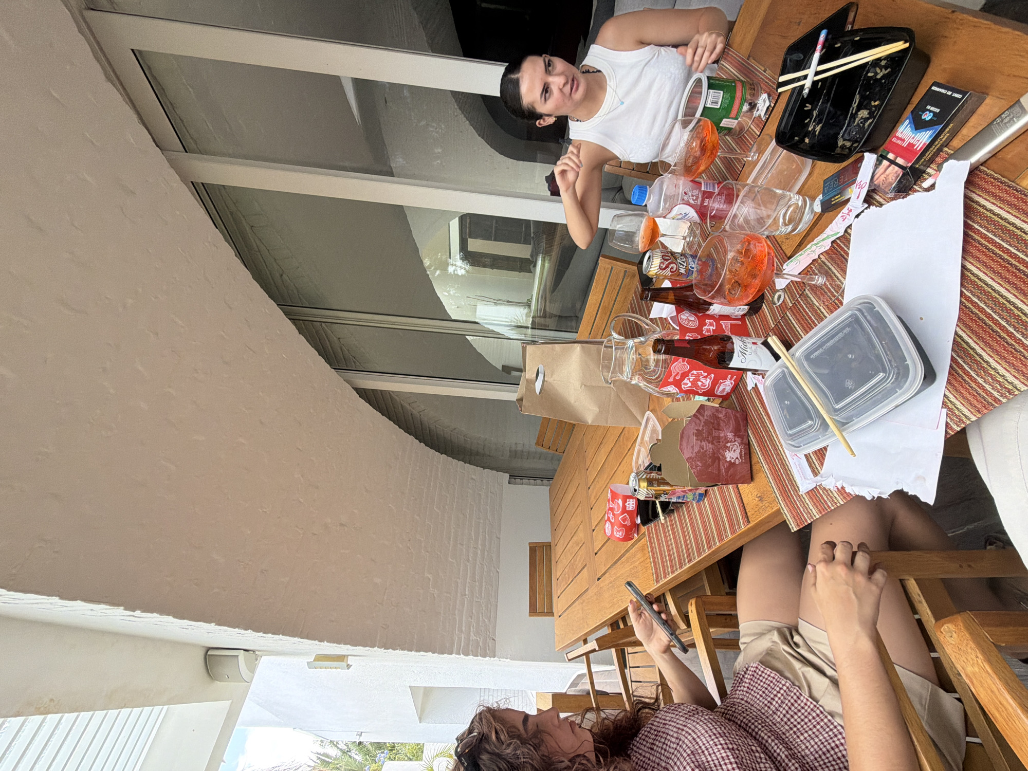 This image captures a casual, outdoor gathering in San Bernardino Tlaxcalancingo, Mexico, showing two women seated at a wooden dining table laden with food and drink remnants. The scene appears to be a patio or balcony, with white textured walls and large windows reflecting interior elements visible in the background.
In the foreground, a woman with dark, curly hair, wearing a plaid shirt and tan shorts, is seen from the chest up, focused on her smartphone. Her left hand holds the phone, while her right hand is casually placed on her leg.
Across the table, another woman with dark hair pulled back, wearing a white tank top and a simple silver necklace, looks towards the camera. She has a neutral expression, with her left arm resting on the table and her right hand slightly raised, as if mid-gesture or thought.
The wooden table between them is covered by two striped placemats in shades of red, white, and brown. It's cluttered with numerous empty or partially filled glasses containing a bright orange liquid (possibly Aperol Spritz), empty beer bottles (one partially visible with "Miche..." text, another "Heineken" can), plastic water bottles, and takeaway food containers. A black plastic tray holds chopsticks and a cigarette butt. Nearby are a red "Marlboro" cigarette pack displaying "CODE 20 GRAMOS," and a white "555" cigarette pack. Several red paper packets with Chinese characters are also scattered on the table, along with crumpled papers and a brown paper bag.
The setting suggests daytime due to the bright, diffused light. The overall atmosphere is relaxed and informal, possibly after a meal with friends.