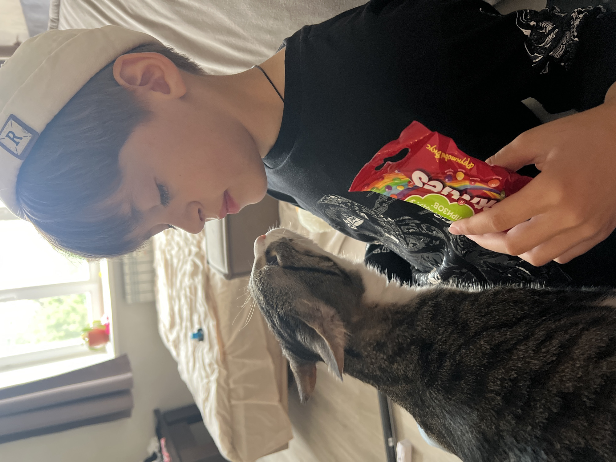 The image captures a close-up indoor scene in the city of Chaikovsky, Russia, featuring a young boy and a domestic cat. A tabby cat, predominantly grey and brown striped with white chest and muzzle, is positioned in the lower-left foreground, looking upwards towards the boy with its ears slightly back. To the right, a young boy is visible from the mid-chest upwards, wearing a cream-colored beanie with a square patch displaying a white 'R' and a black t-shirt. His eyes are closed or directed downwards. He holds a red bag of Skittles candy, marked with "Фруктовый Вкус" (Fruit Flavor) and "ПРИЗОВ" (Prizes) in Cyrillic, in his left hand. A thin dark cord necklace is around his neck. The background reveals an unmade bed with a light-colored duvet, upon which a small blue object rests. To the left, a partially visible dark bedside table stands beneath a window with light-brown curtains, through which bright natural light and green foliage are visible. A white radiator is present below the window. The floor is a light-toned wooden or laminate surface.