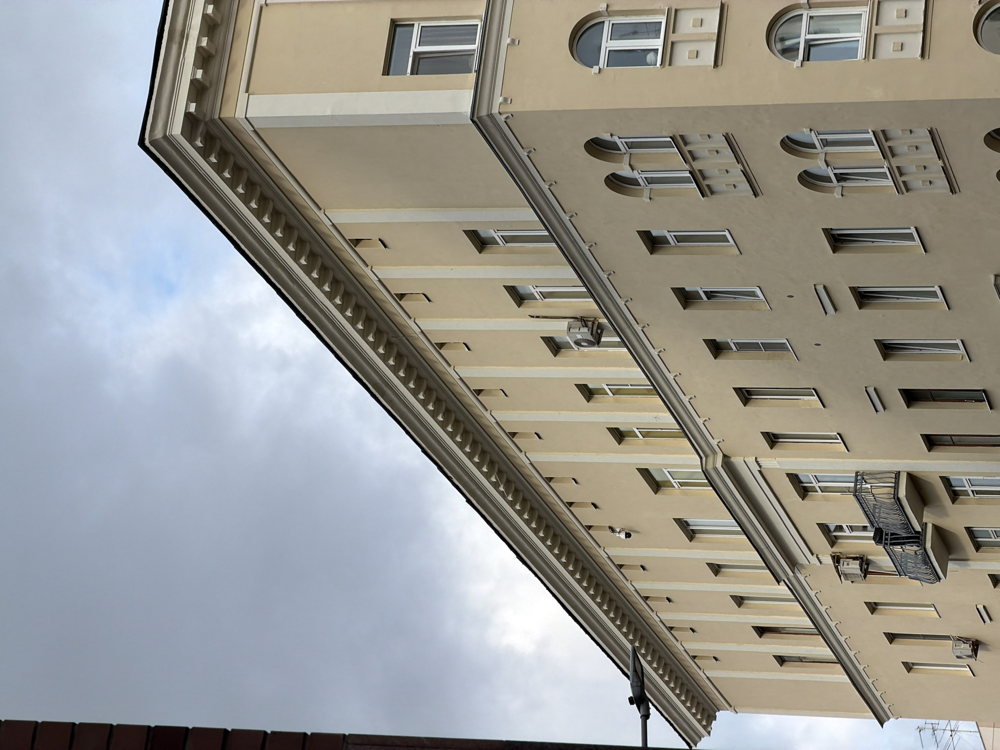 The image shows the facade of a light yellow, multi-story building in Moscow, Russia. The building features a decorative cornice along the top and architectural vertical elements between the windows. Numerous windows are visible, some rectangular and some arched, with white frames. Several air conditioning units are mounted on the exterior walls. A small balcony with a wrought-iron railing is present on the second floor. The sky above is overcast with clouds. A section of a red brick building is partially visible on the left edge of the frame, along with a street lamp. There are no people or vehicles clearly visible in the image.