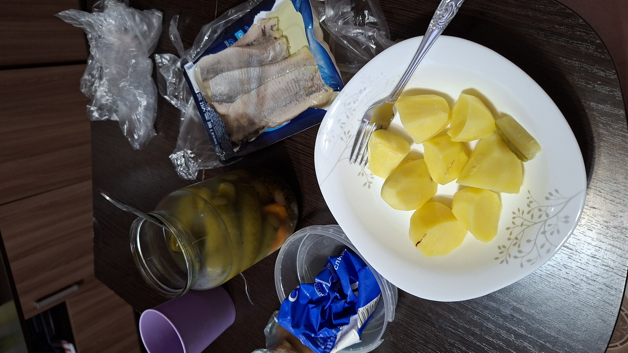 The image shows a dark wooden table with food and packaging. In the foreground, a white plate with a floral design holds several pieces of peeled, boiled potatoes. A fork rests on the plate, positioned to the right of the potatoes. To the left of the plate is a clear plastic container with a crumpled blue wrapper inside. Behind these items, a glass jar filled with pickles and brine is visible, with a spoon sticking out of it. Further back and to the right, a plastic-wrapped package of herring fillets is placed on the table, alongside crumpled plastic bags. A purple cup is partially visible on the far left side of the image. The scene suggests a meal being prepared or consumed. The background appears to be a domestic setting, possibly a kitchen or dining area, with wooden cabinetry visible. The image does not contain any specific clues to pinpoint its location within Novoilinsky, Russia.