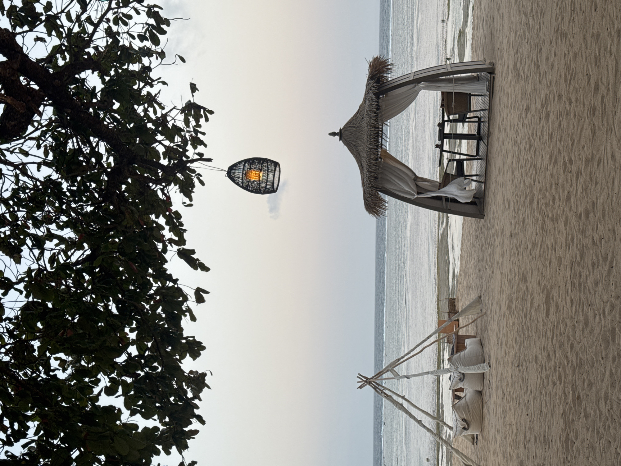 The image depicts a serene beach scene in Kuta Selatan, Indonesia, under a dusky sky. In the foreground, a sandy beach stretches towards the gentle ocean. To the left, a triangular tent-like structure, constructed with wooden poles and draped with light fabric, houses several large, white beanbag chairs and a low table. To the right, a more elaborate open-air cabana with a thatched roof and flowing white curtains offers a dining setup, complete with a table and two chairs. Above the cabana, a decorative woven lantern hangs from a tree branch, its interior light glowing. The ocean in the background is calm, with visible ripples and a distant horizon. The overall atmosphere is tranquil and inviting, suggesting a peaceful evening on the coast.