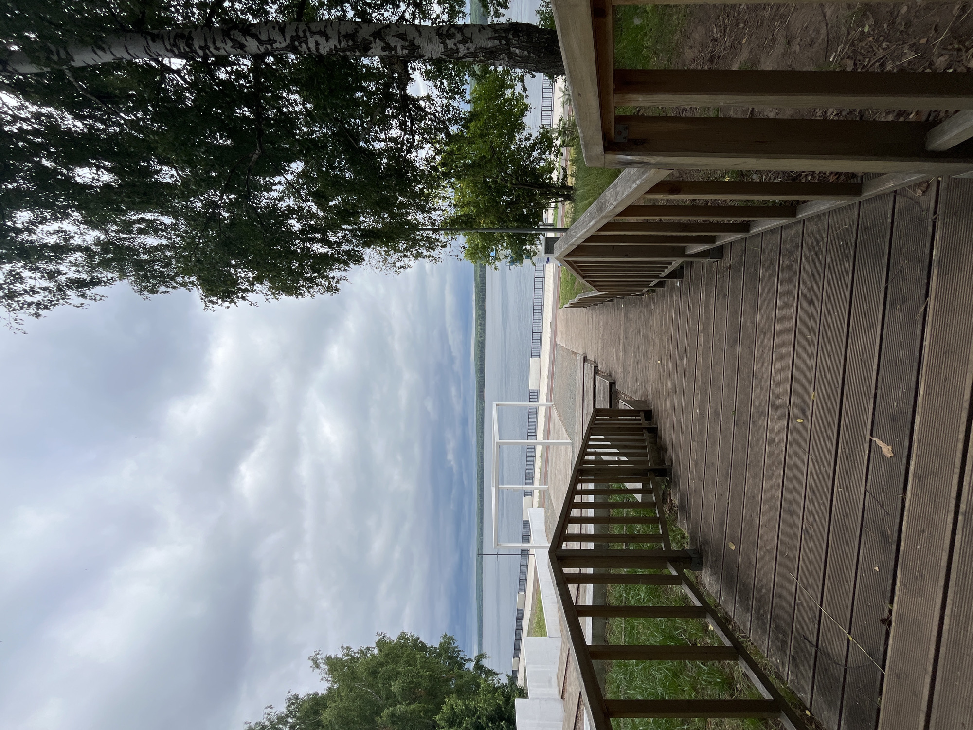 The image depicts a wooden staircase leading down towards a waterfront area in the city of Chaikovsky, Russia. The sky is overcast with thick, gray clouds, suggesting a temperate or cooler climate.
In the foreground, the focus is on the weathered wooden staircase with sturdy railings on both sides. The wood appears aged and shows signs of outdoor exposure, with some leaves scattered on its surface. The staircase is designed with wide steps, and it curves slightly as it descends.
Beyond the staircase, the scene opens up to a paved promenade along a wide body of water, likely a river or a reservoir given the location. The water is calm and reflects the muted tones of the sky. A white, modern structure, resembling a canopy or gazebo, stands on the promenade, offering a stark contrast to the natural surroundings and the rustic staircase. White concrete walls are also visible on the left side of the promenade, partially framing the view.
Green trees, including a prominent birch tree on the right with its distinctive white bark, flank the scene, adding a natural element to the urban waterfront. The background reveals a distant treeline and hills on the opposite shore, emphasizing the vastness of the water. The overall atmosphere is peaceful and somewhat somber due to the cloudy weather, highlighting a public recreational space in Chaikovsky.