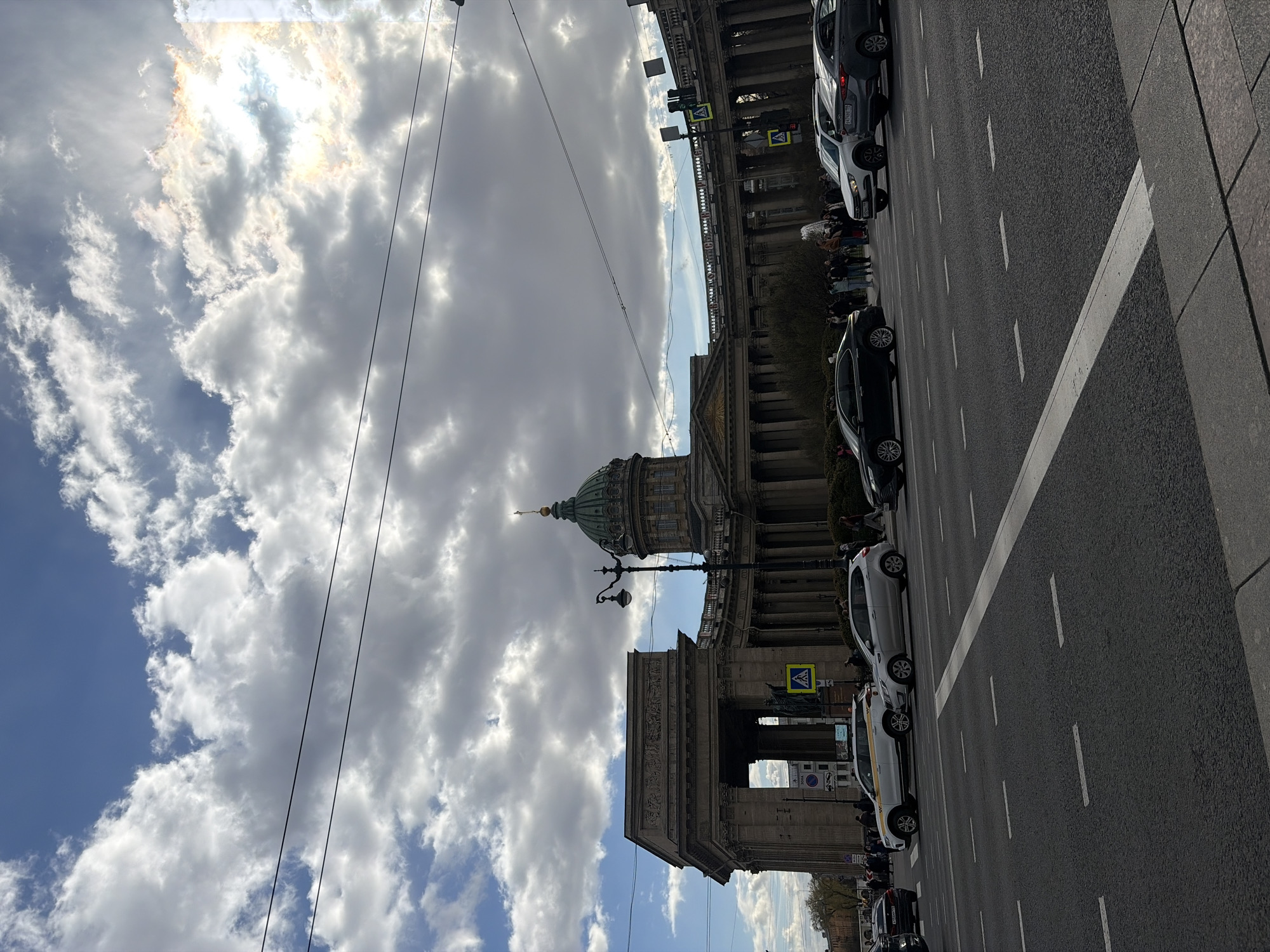 The image displays a bustling city street scene, dominated by the Kazan Cathedral in Saint Petersburg, Russia, situated on the left side of the frame. The cathedral is a grand neoclassical building characterized by its extensive colonnade and a distinctive green dome topped with a golden spire.
In the foreground, a wide, multi-lane asphalt road runs parallel to the cathedral. White dashed lines mark the lanes, and a solid white line defines the edge of the road next to a sidewalk made of grey paving stones. Several cars are parked along the right side of the street, including vehicles of white, dark grey, and black colors. People are visible walking on the sidewalk next to the parked cars and in front of the cathedral.
The sky fills a large portion of the upper image, featuring a dynamic mix of bright blue patches and numerous fluffy white and grey clouds. The sun is shining brightly from behind some clouds, creating a brilliant glow. Overhead power lines stretch across the sky. An ornate black lamppost stands in the mid-ground, providing street illumination. Yellow pedestrian crossing signs (diamond-shaped with a black walking figure) are visible on poles near the parked cars. Some car license plates are visible, consistent with Russian format. The scene depicts a bright, daytime setting.