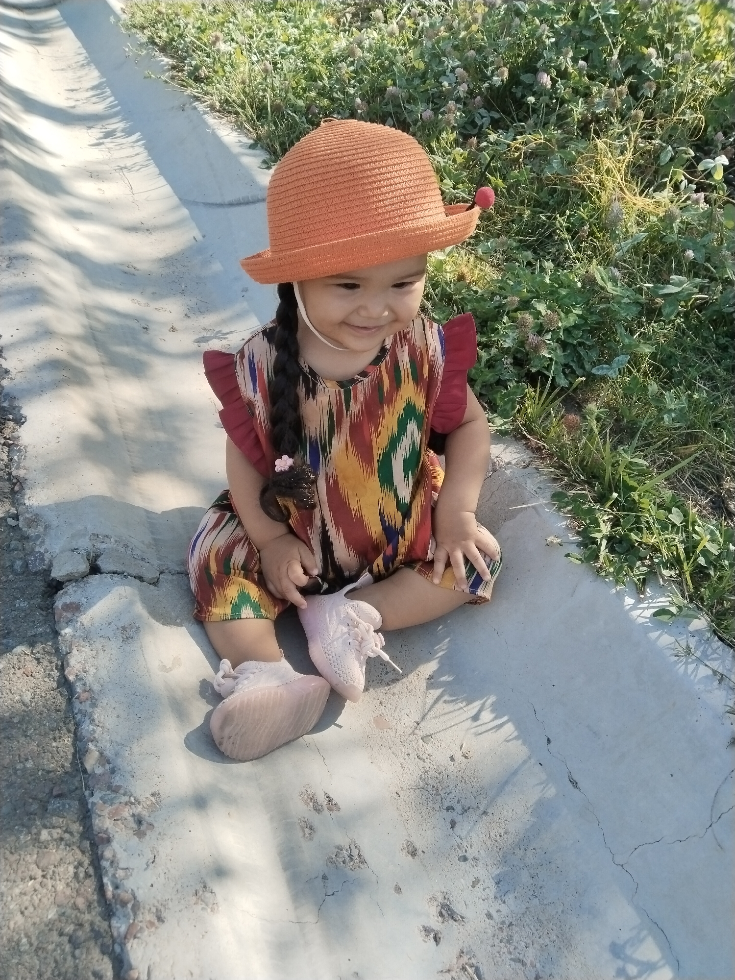 A young child wearing an orange hat and a colorful patterned dress sits on a concrete surface next to a grassy area in Akata, Uzbekistan. The child has dark hair in a braid adorned with a pink flower. The child's feet are clad in pink sneakers. The concrete area appears to be a shallow ditch or channel, with shadows cast by vegetation. The background is filled with lush green grass and some small plants with pinkish-purple flowers. The scene is illuminated by sunlight, creating dappled shadows.