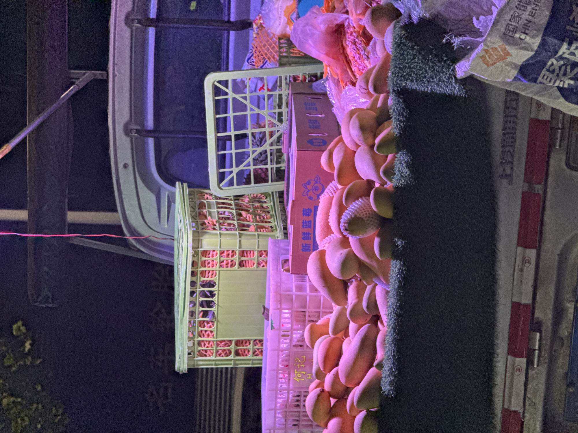 The image captures a scene at night in the Yuzhong District, China, featuring an overloaded vehicle. Crates of strawberries and a mountain of mangoes are stacked precariously on the vehicle. The boxes carrying the mangoes have Chinese characters. Other objects include a white plastic crate and some packaging for an "上汽通用五“ car tire. The environment is dark, suggesting an outdoor setting during the nighttime. The overall impression is of a mobile fruit stand or market, with goods spilling out of the vehicle.