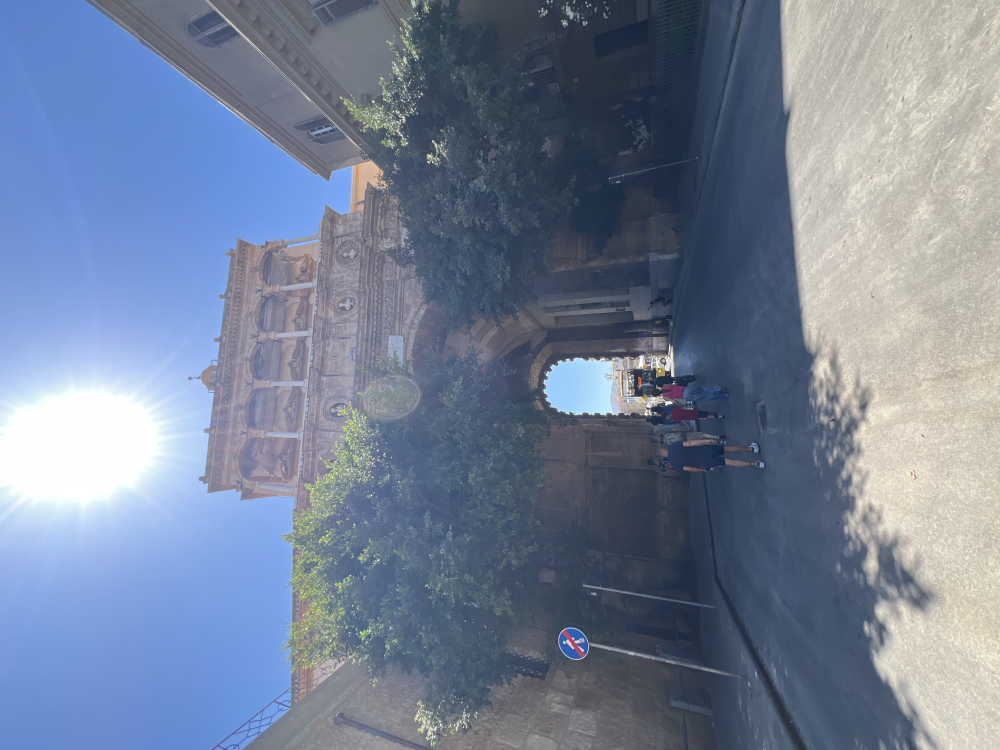 The image depicts a street scene in Palermo, Italy, on a bright, sunny day. The sun is high in the sky, casting sharp shadows. The main subject is a grand, ornate stone archway that appears to be part of an ancient building, possibly a historic monument or entrance. Lush green trees grow around and through the archway, creating a natural contrast with the stone architecture.
A group of people are walking through the archway and along the paved street. They are casually dressed, suggesting a typical day of exploration or daily life. The perspective is from street level, looking towards the archway and the bright sky beyond.
Notable details include the intricate carvings and architectural elements of the stone structure. A circular sign with a red border and a horizontal red bar across a symbol, likely indicating a restriction, is visible on a pole beside the street. The overall mood is one of vibrant activity and historical charm. The time of day appears to be midday, given the intensity of the sunlight and the length of the shadows. The location is identifiable as Palermo, Italy, contributing to the sense of cultural richness and historical depth. No visible text is discernible in the image.