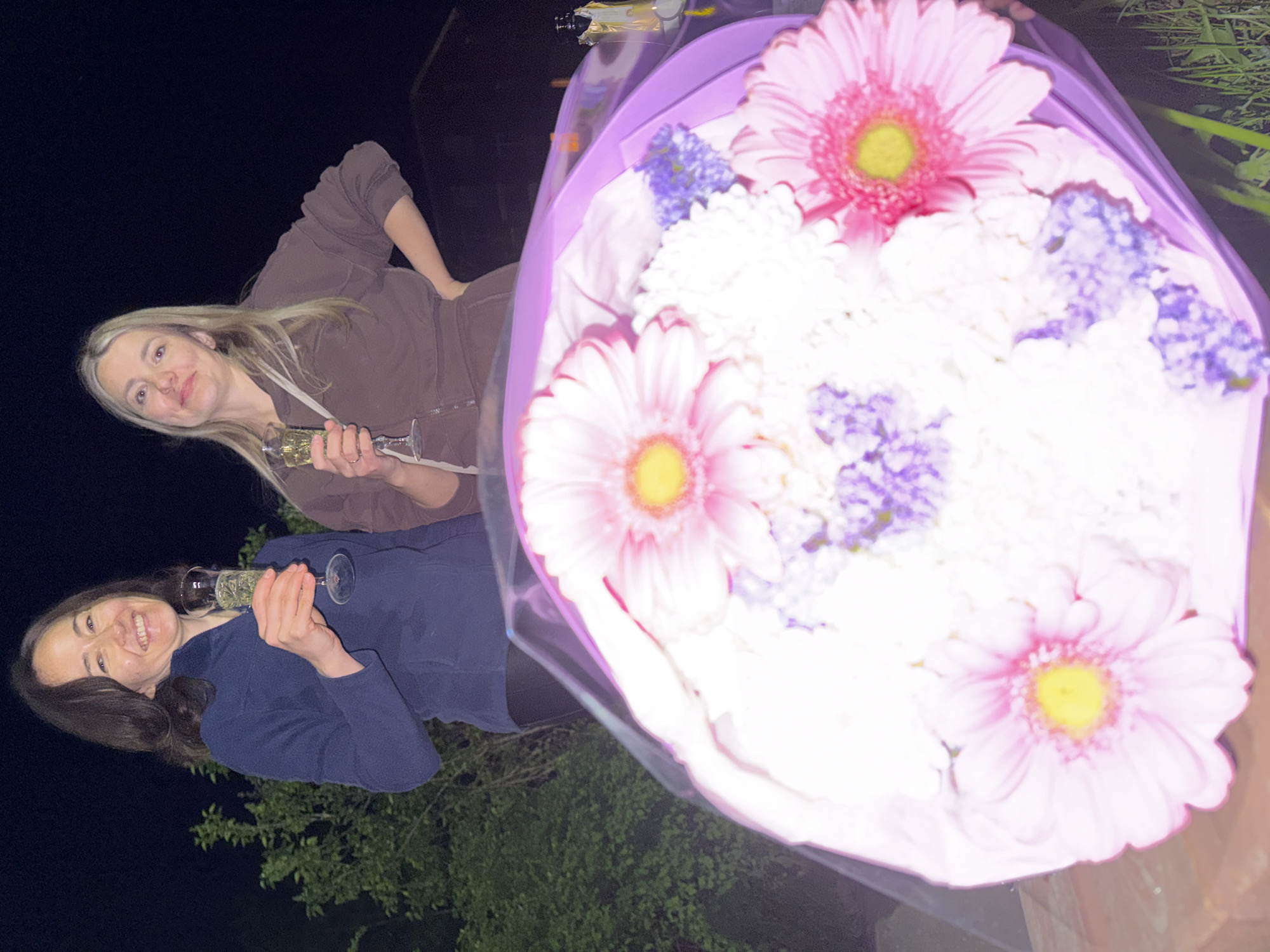 Two women are photographed at night, holding champagne flutes and a bouquet of flowers. One woman, with brown hair, wears a navy blue sweater. The other, with blonde hair, is in a brown sweatsuit. The bouquet contains pink gerbera daisies, white chrysanthemums, and purple flowers, wrapped in purple paper. The background is dark, with some foliage visible. No location in Votkinsk, Russia can be ascertained from this image.