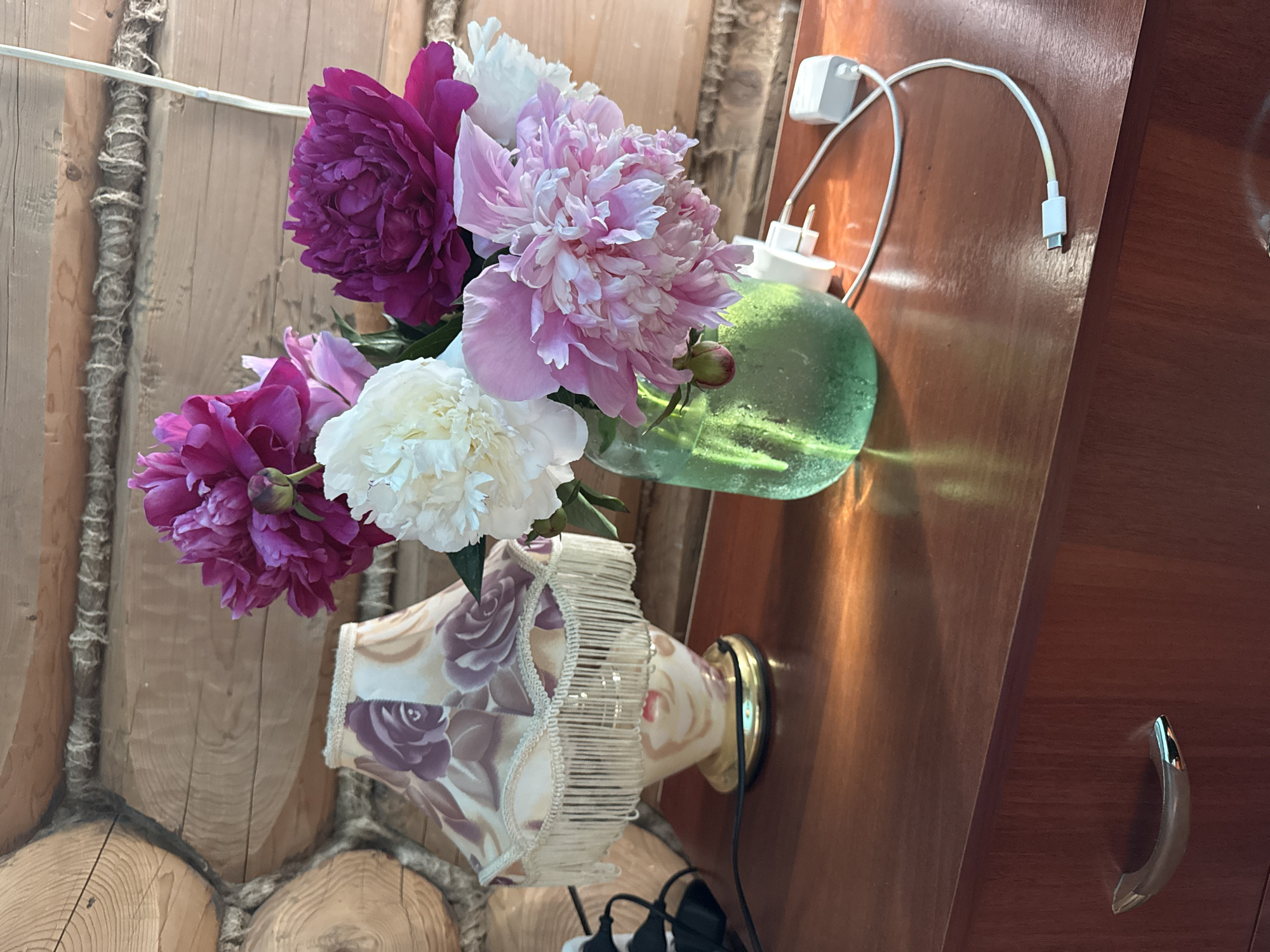 A bouquet of pink and white peonies is arranged in a green glass jar on a wooden dresser. The jar is to the right of a fringed table lamp with a floral pattern. The dresser has a wooden handle on the drawer. The background is a log wall. A white charging cable is plugged into a white adapter on the dresser. The setting appears to be a room in Чуй-Атасево, Russia.