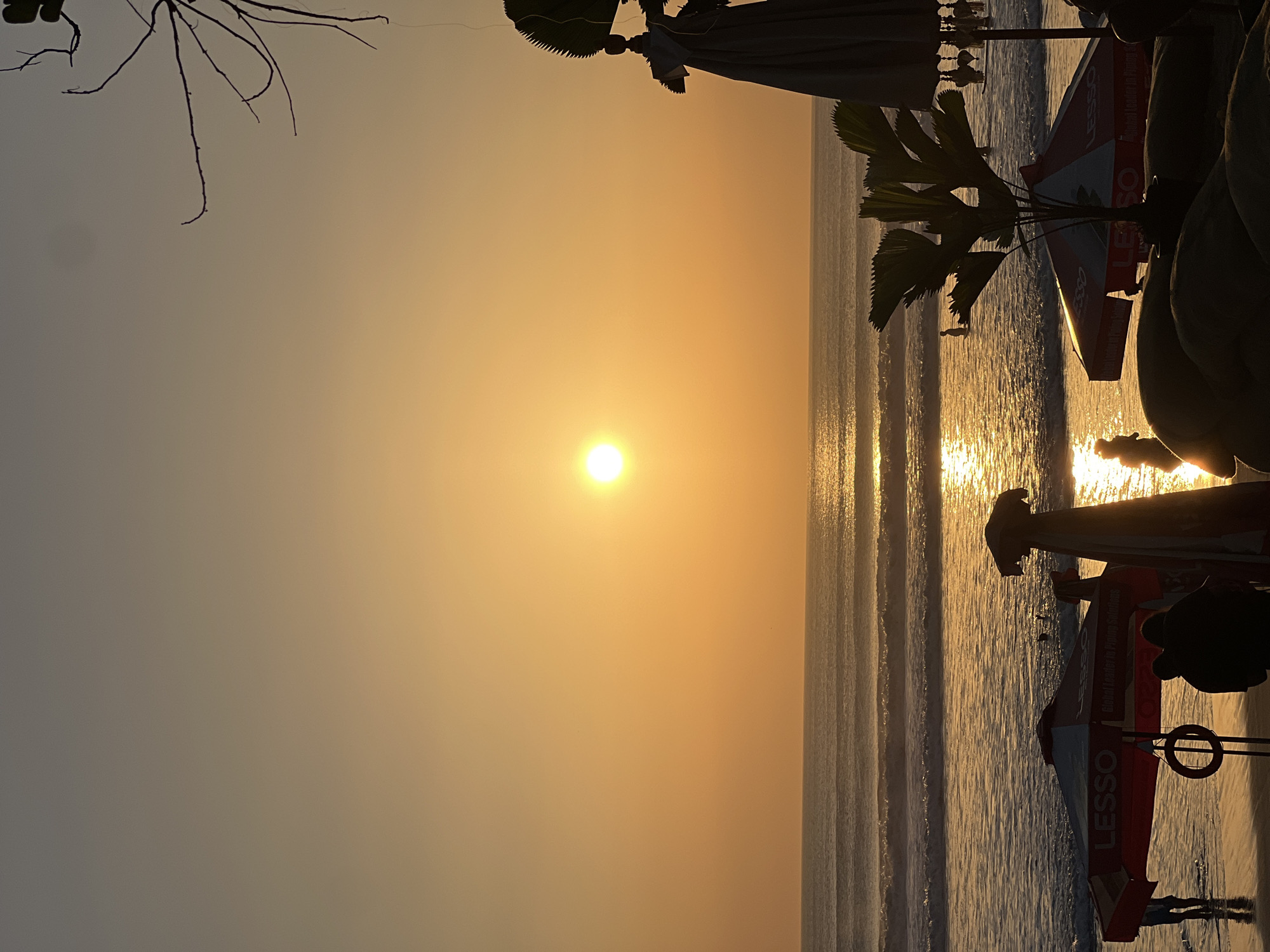 The image depicts a sunset scene at Kuta Beach, Bali, Indonesia. The sun is setting over the ocean, casting a warm, golden glow across the sky and water. The ocean has gentle waves breaking on the shore. In the foreground, there are several beach umbrellas, some with the "LESSO" logo visible, and beachgoers can be seen in the distance wading in the water and walking on the sand. Silhouetted against the bright sky and ocean are palm trees and other beachside structures. The overall atmosphere is tranquil and picturesque, capturing the essence of a Balinese sunset.