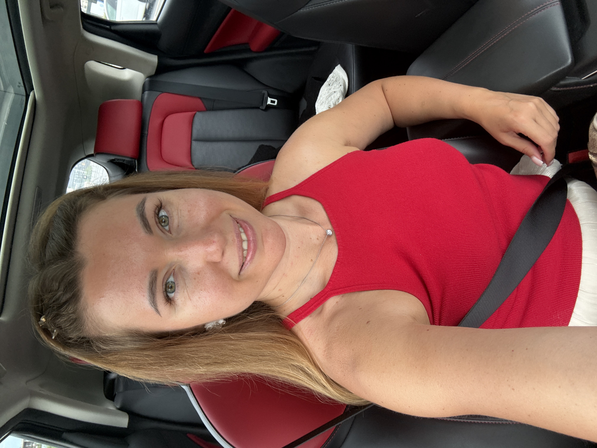 A woman with long blonde hair is taking a selfie in a car. She is wearing a red tank top and a silver necklace with a small square pendant. She is sitting in the passenger seat, and the camera is positioned to capture her from the chest up. The car has black and red leather seats. The background shows the interior of the car, including the headrests and a portion of the sunroof. There is a seatbelt visible across her chest.