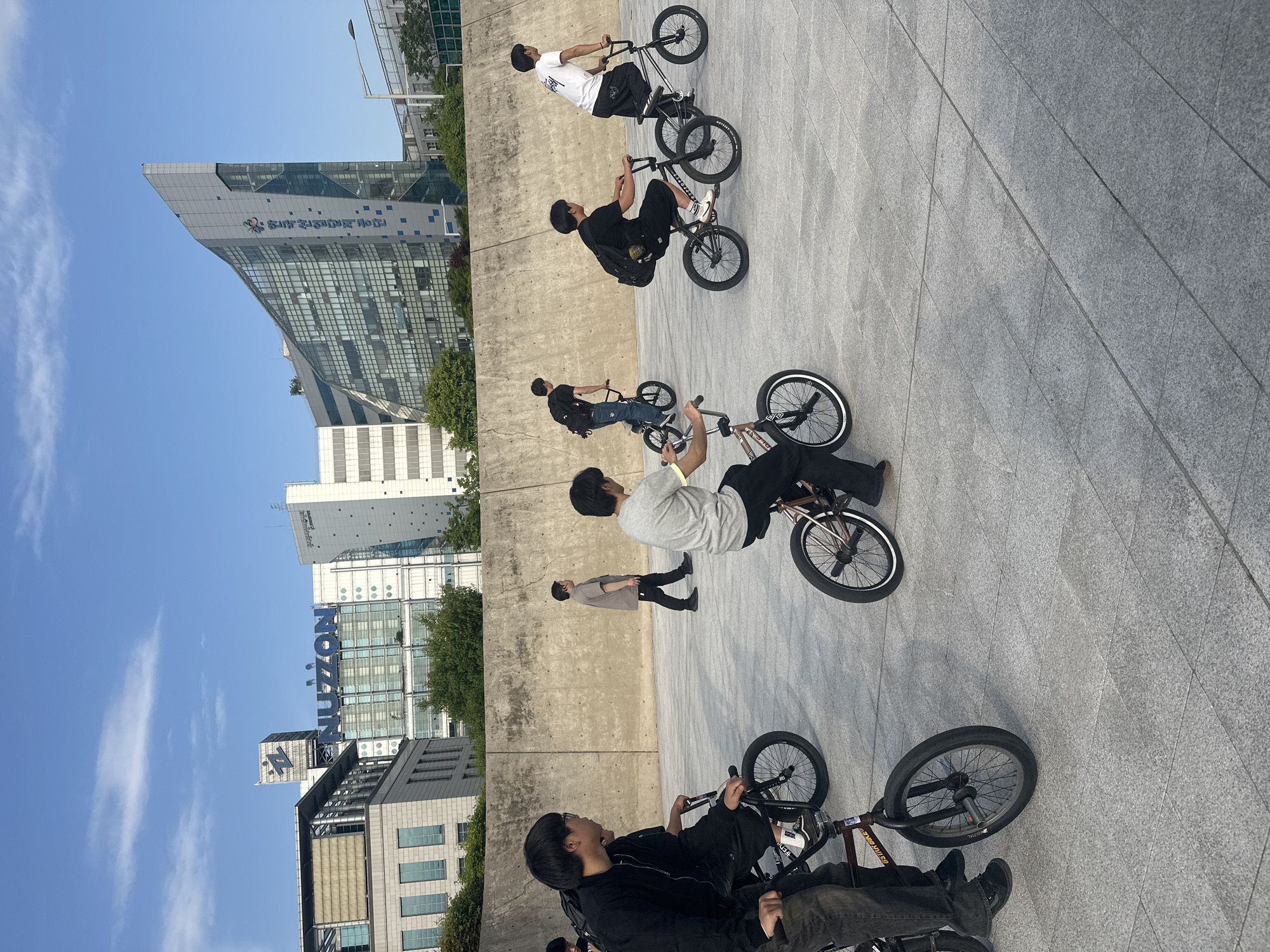A group of six people are in a plaza or large paved area in Seoul, South Korea. Five individuals are on BMX bicycles, with varying positions and stances on the bikes. One person is walking. The area is bordered by a wall, and behind the wall are modern buildings. NUON and another logo are visible on one building. The sky is blue with some clouds. The image is rotated.