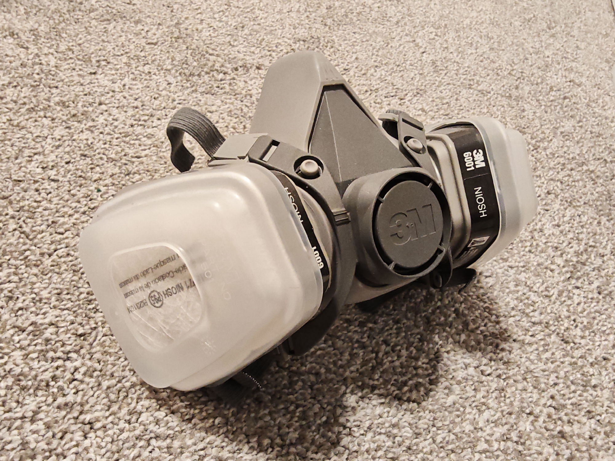 A close-up, slightly angled, high-angle shot shows a gray and white 3M respirator with a clear plastic cover over one of its filters, resting on a textured, light brown carpet. The respirator is positioned with its front facing slightly left. The main body of the respirator is gray, with a black strap visible at the top left. The clear plastic cover has white text printed on it, including "771 NIOSH (P95) B2317CN" and "lado-Costado de la masara-masque-lado de la mascara". On the side of the respirator, a black band has white lettering that reads "3M 6001 NIOSH". The background is entirely composed of the carpet. There are no people or discernible geographical markers in the image to indicate its location within Calgary, Canada.